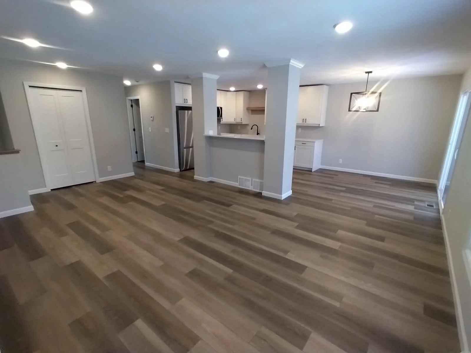 Empty living room with hardwood floors, white walls, recessed lighting, and an open kitchen with white cabinets and stainless steel appliances.