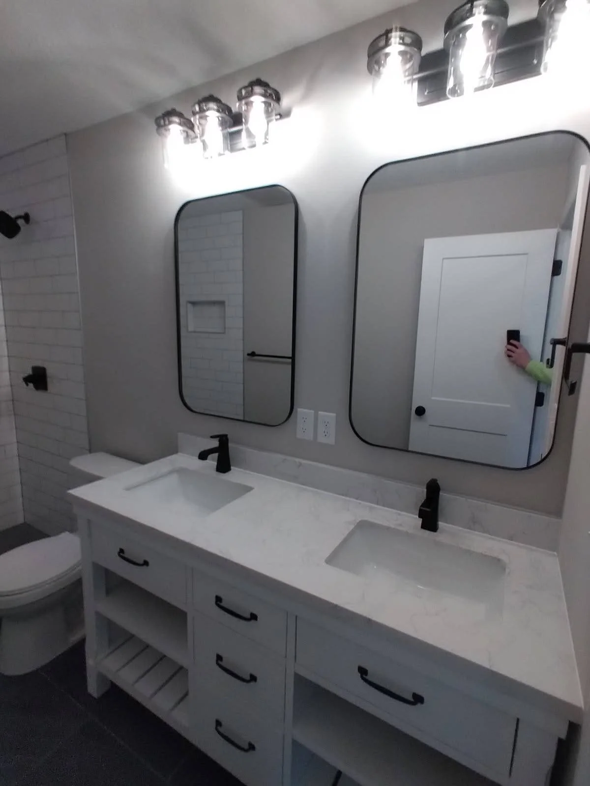 A modern bathroom with a double vanity with black fixtures, two rectangular mirrors, and a white countertop, with a white door and a brick wall visible in the reflection.