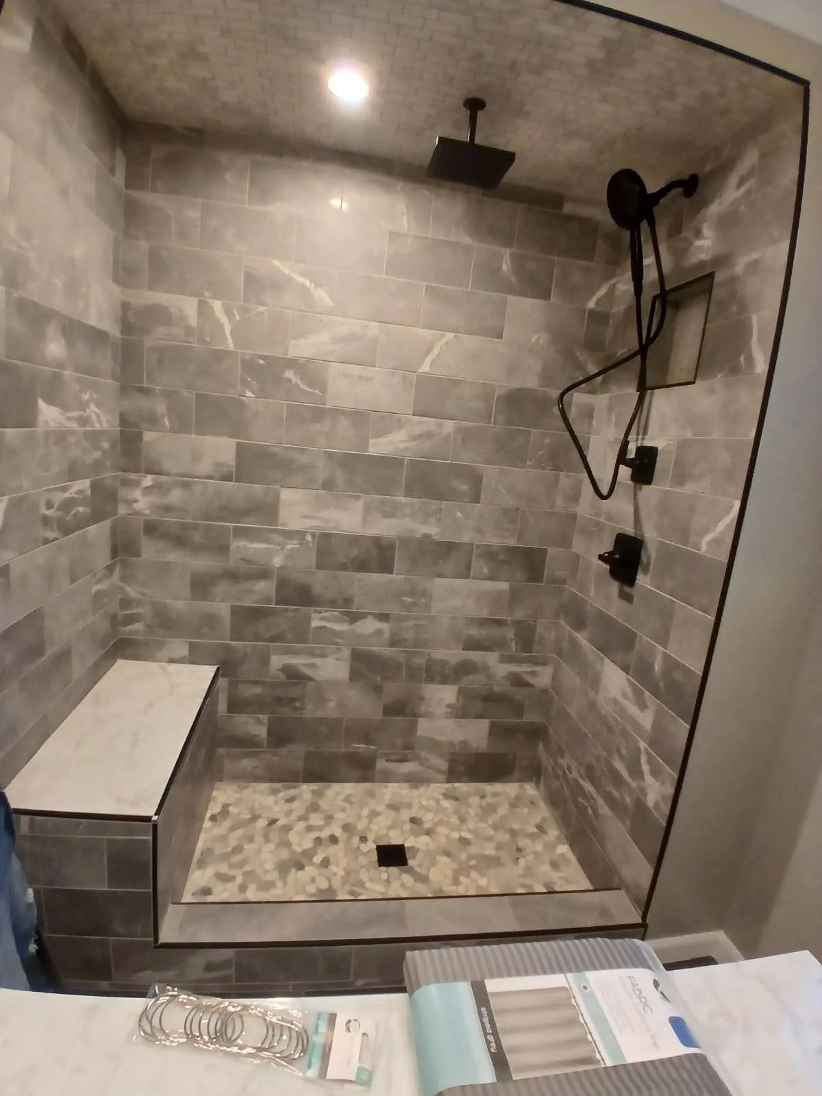 A walk-in shower with gray stone tile walls, a pebble tile floor, a built-in bench, a ceiling-mounted rainfall showerhead, and a handheld showerhead, with a small recessed shelf in the wall.