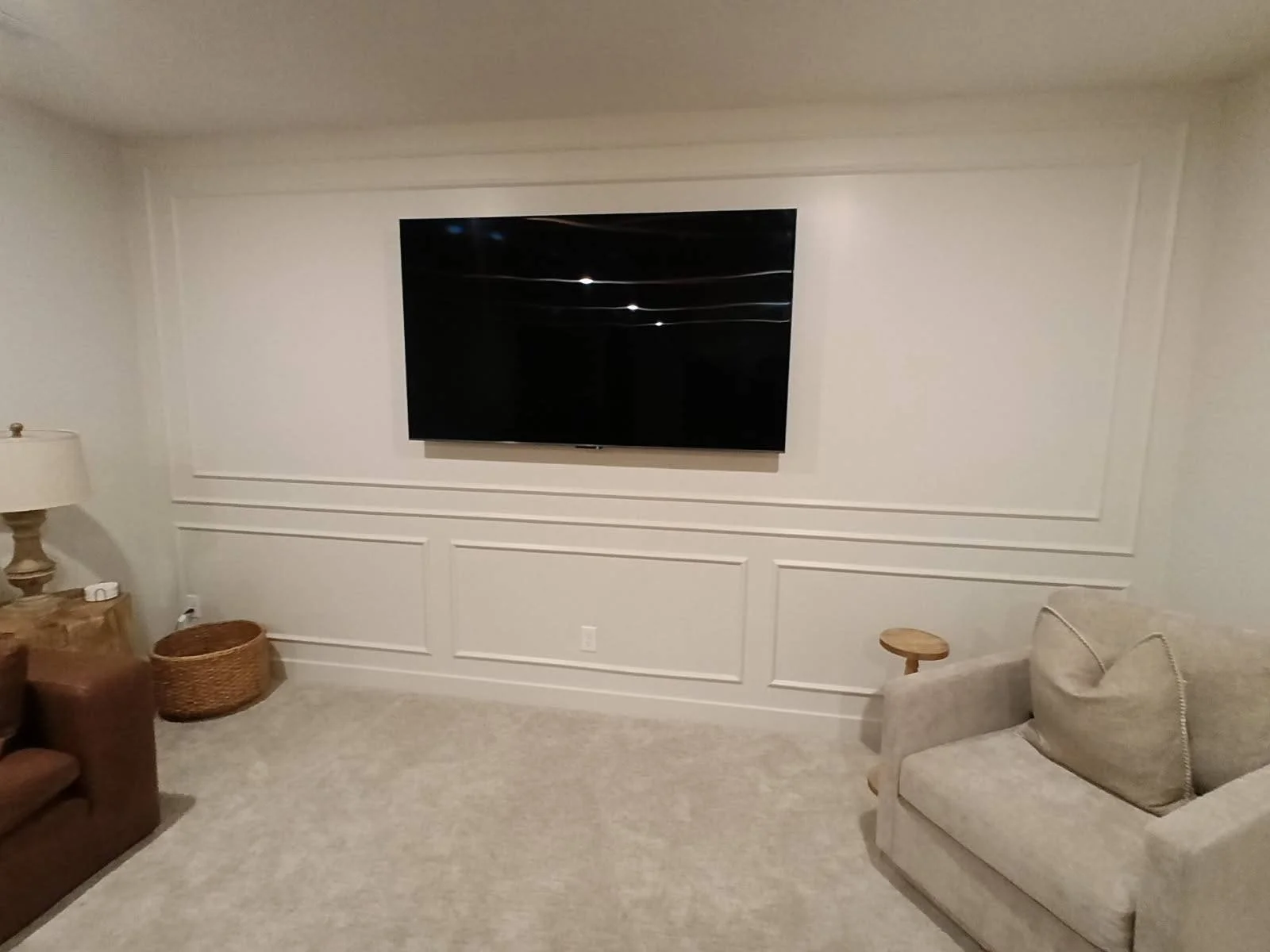 Living room with a wall-mounted flat-screen TV, beige carpet, beige armchair with a pillow, wooden side tables, and a woven basket.