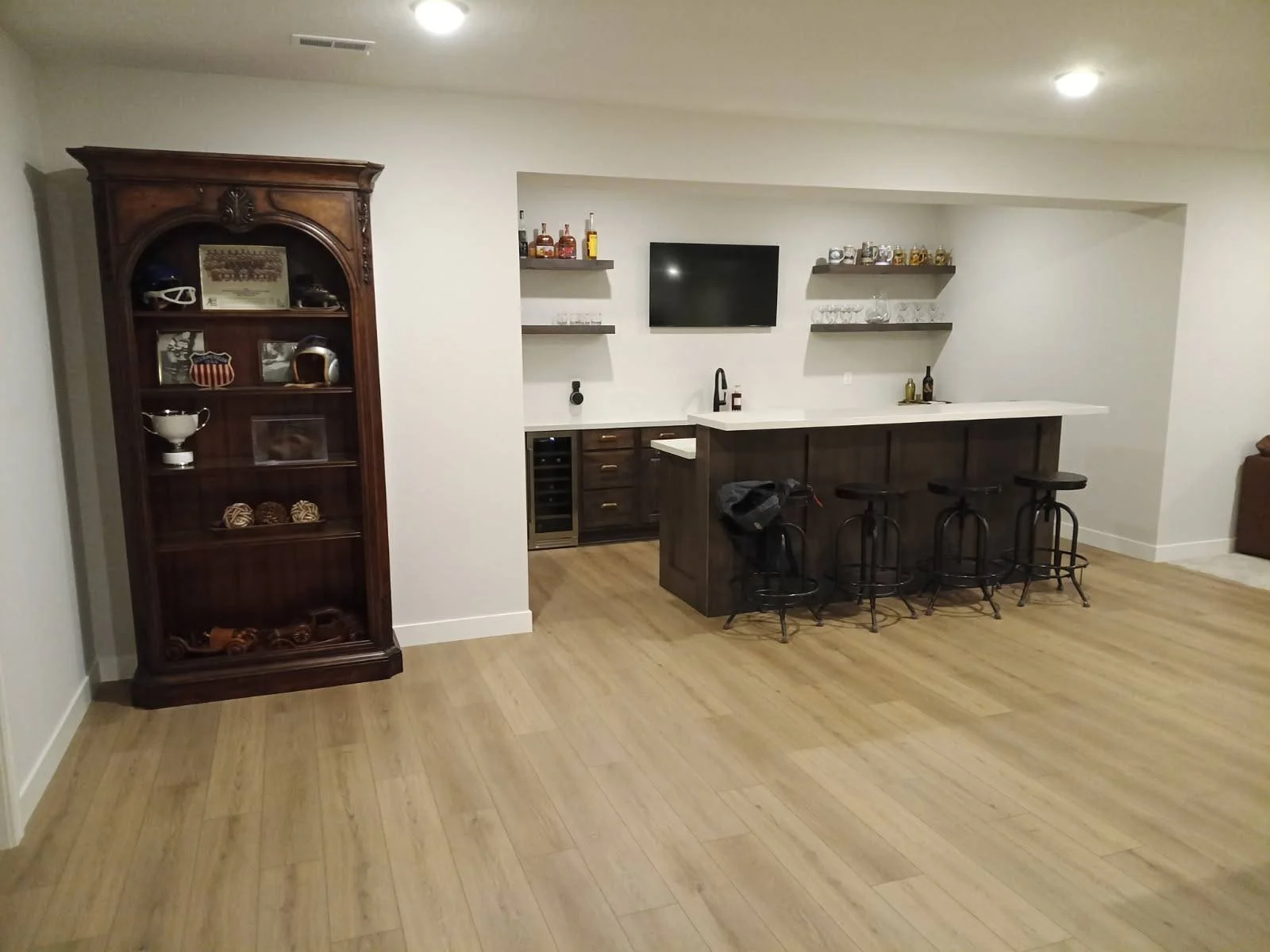 A home bar area with a wooden bar counter, three black bar stools, a wall-mounted TV, shelves with bottles and glasses, and a wooden cabinet with decorative items.
