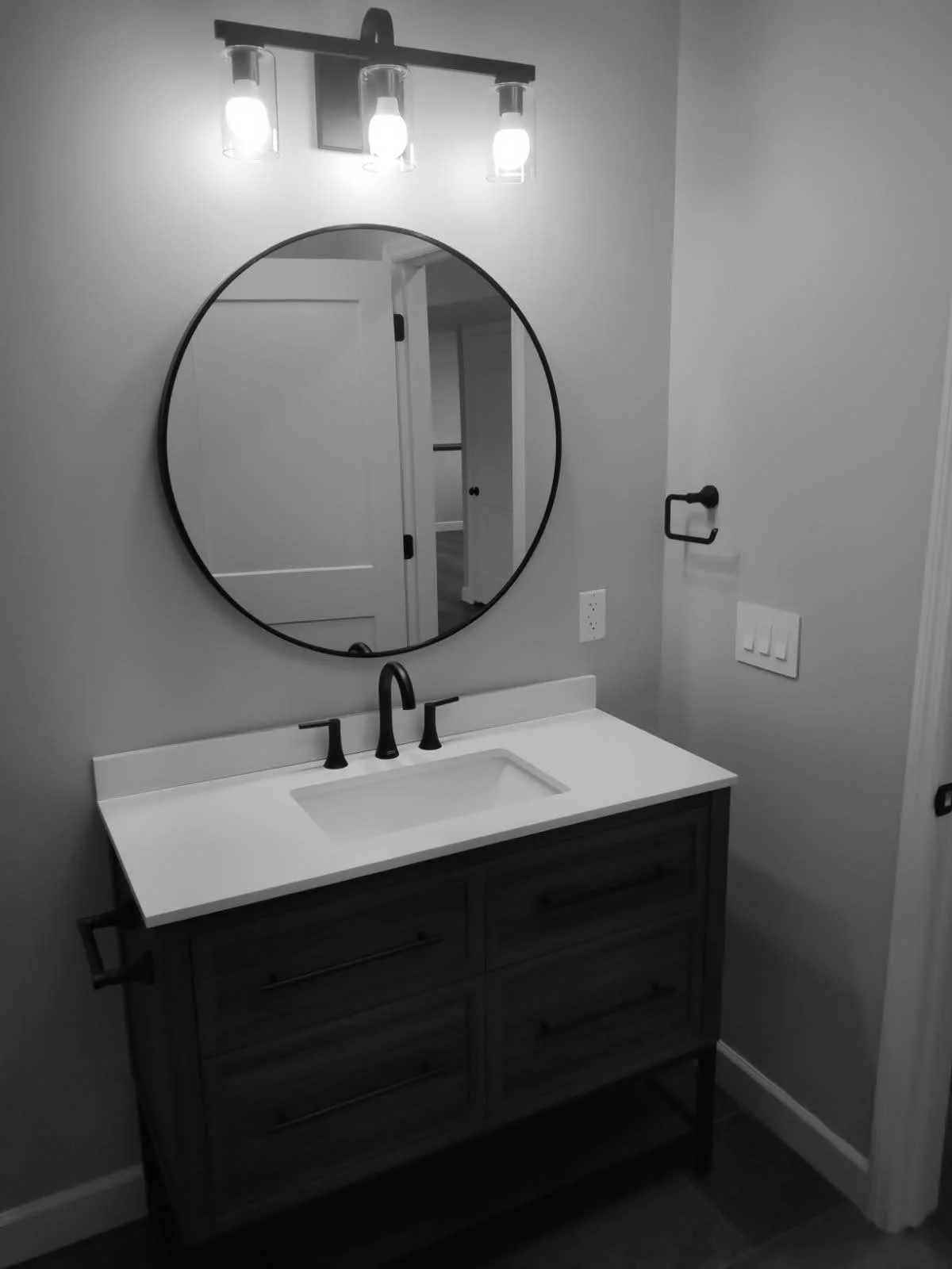 Bathroom vanity with a round mirror, black faucet, and lighting fixture with three bulbs.