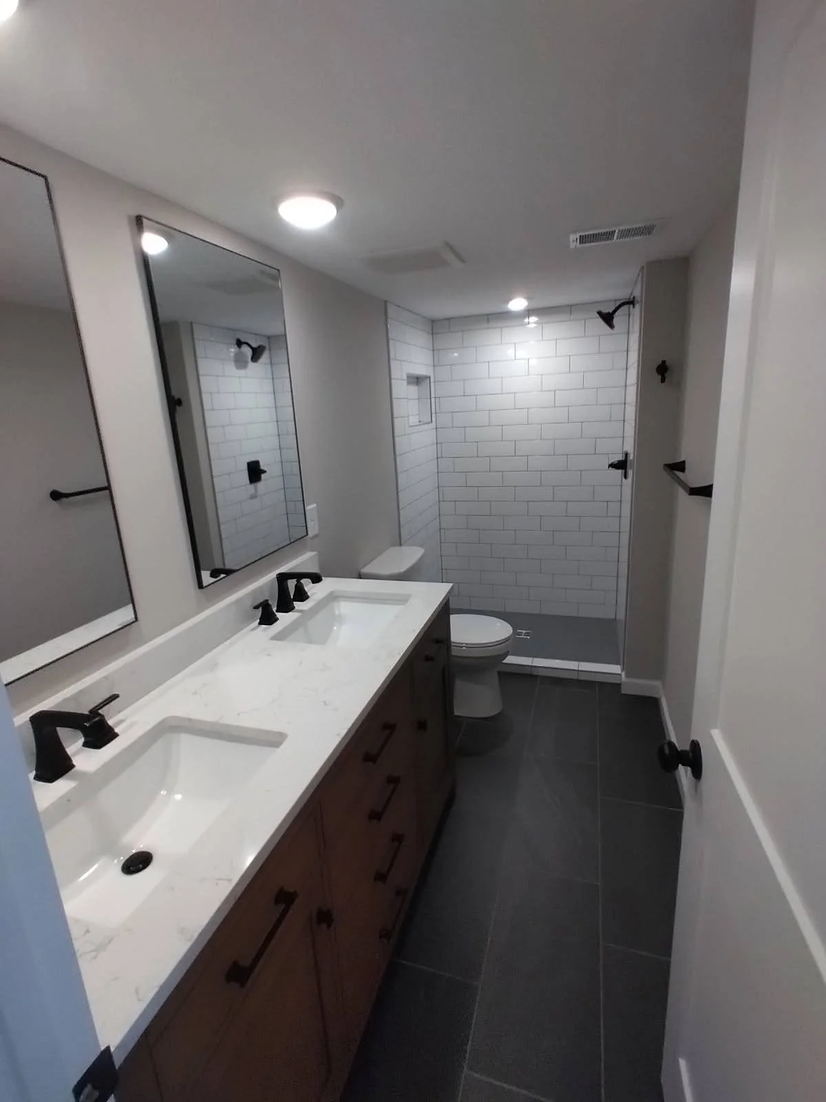Modern bathroom with dual sinks, black faucets, large mirror, white subway-tile shower area with built-in niche, dark floor tiles, and neutral wall paint.