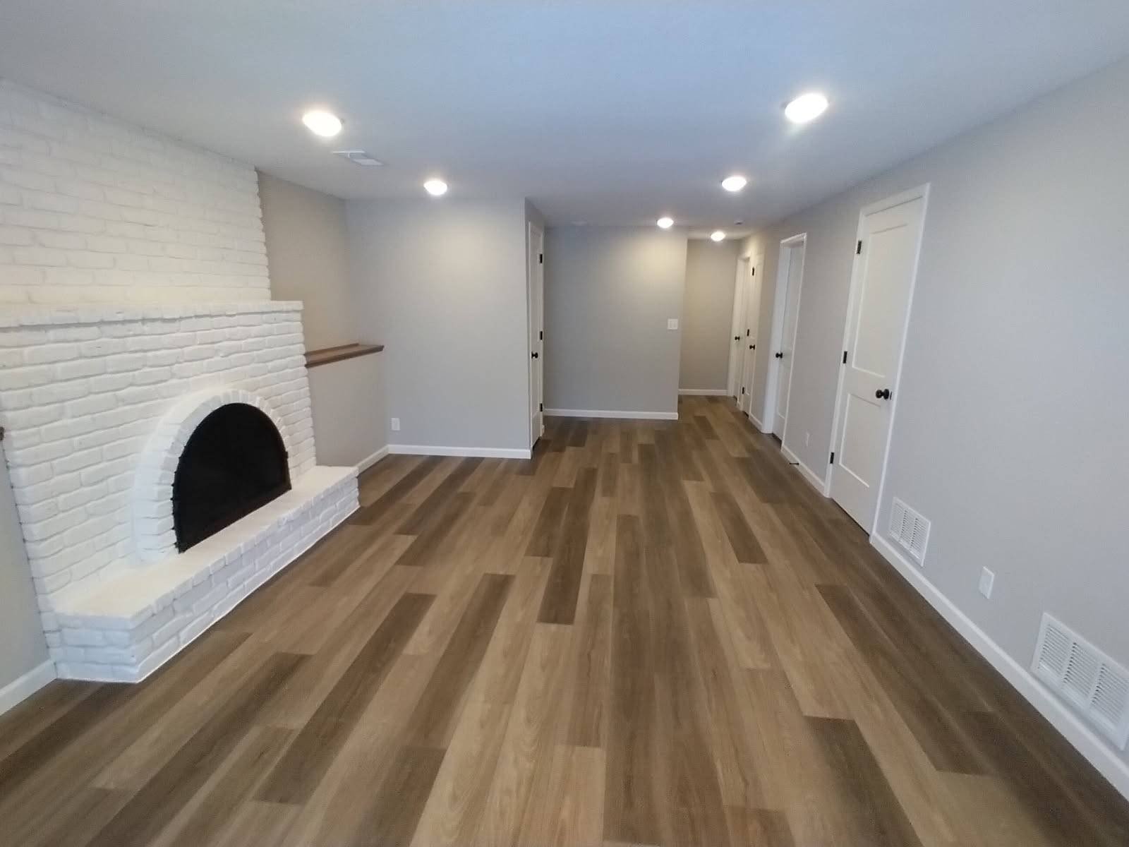 Empty living room with wood flooring, white brick fireplace, white walls, and multiple ceiling lights, white doors, and vents along the base of the walls.