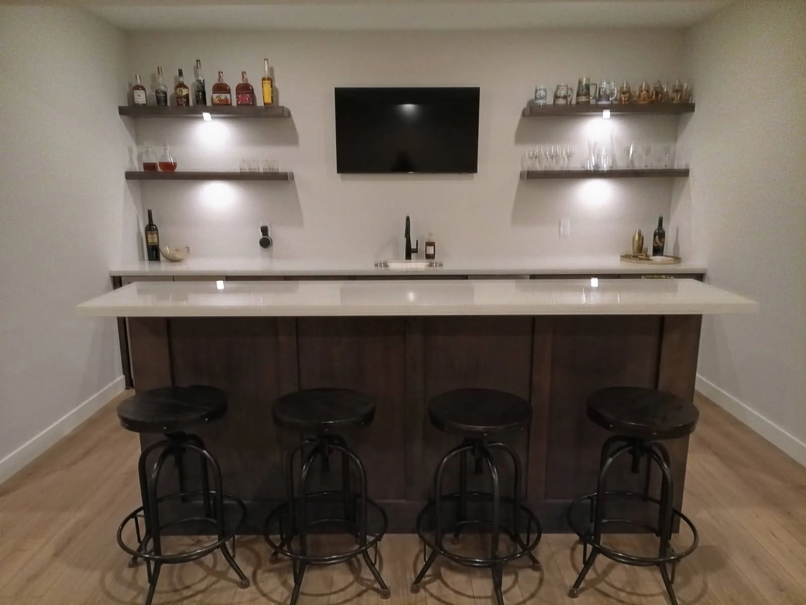 Home bar with four black stools, a white countertop, wall-mounted shelves displaying liquor bottles and glasses, a mounted TV, and a sink in the center.
