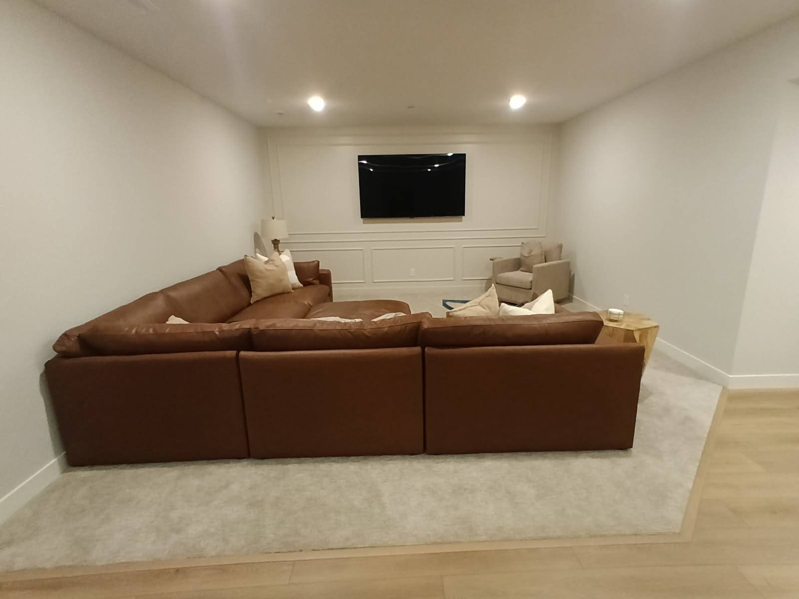 Living room with a brown sectional sofa, beige armchair, and a wall-mounted TV in a minimalist space.