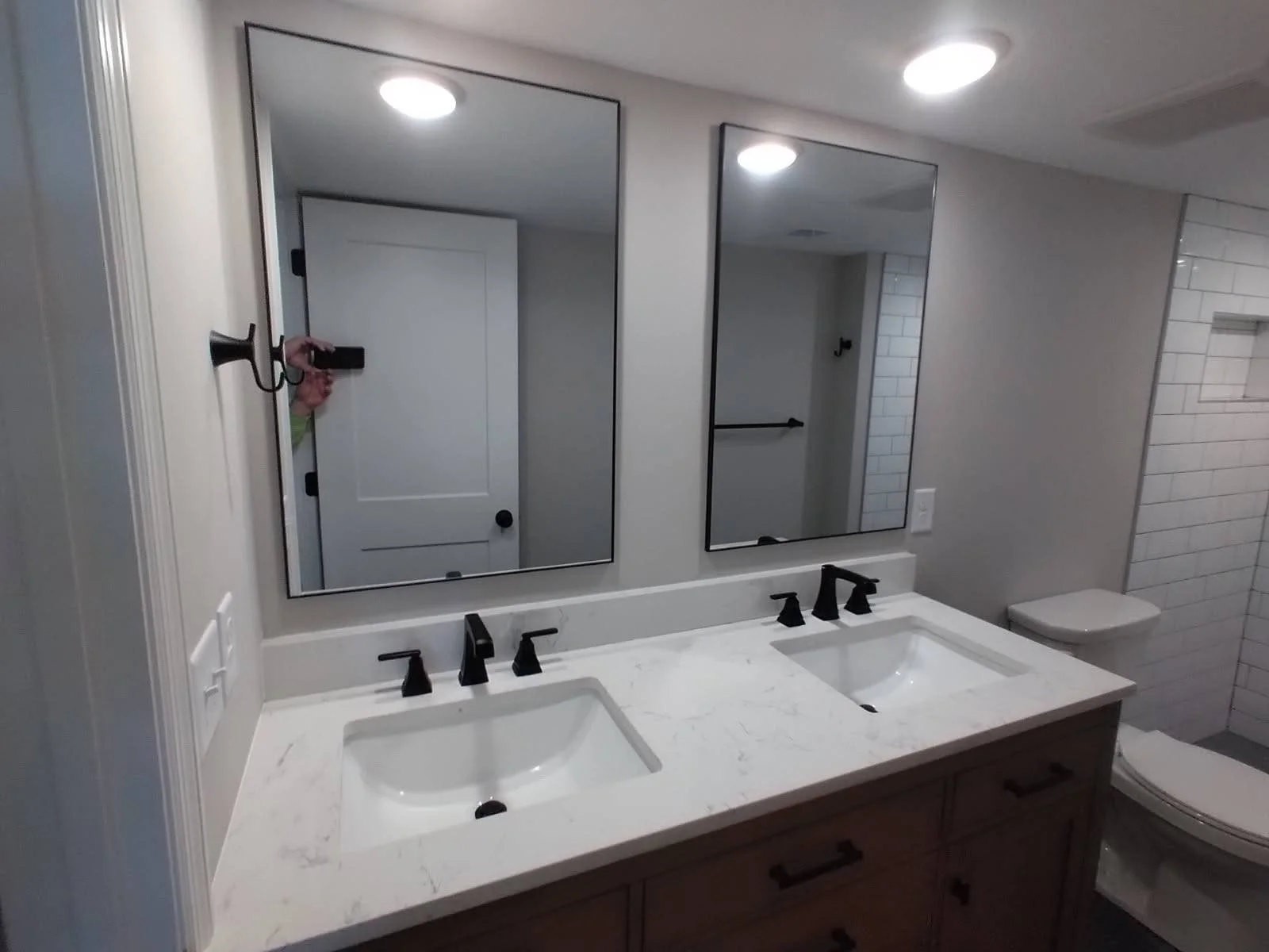 Bathroom with double vanity sinks, two mirrors, black fixtures, white marble countertop, white tiled walls, and a toilet.