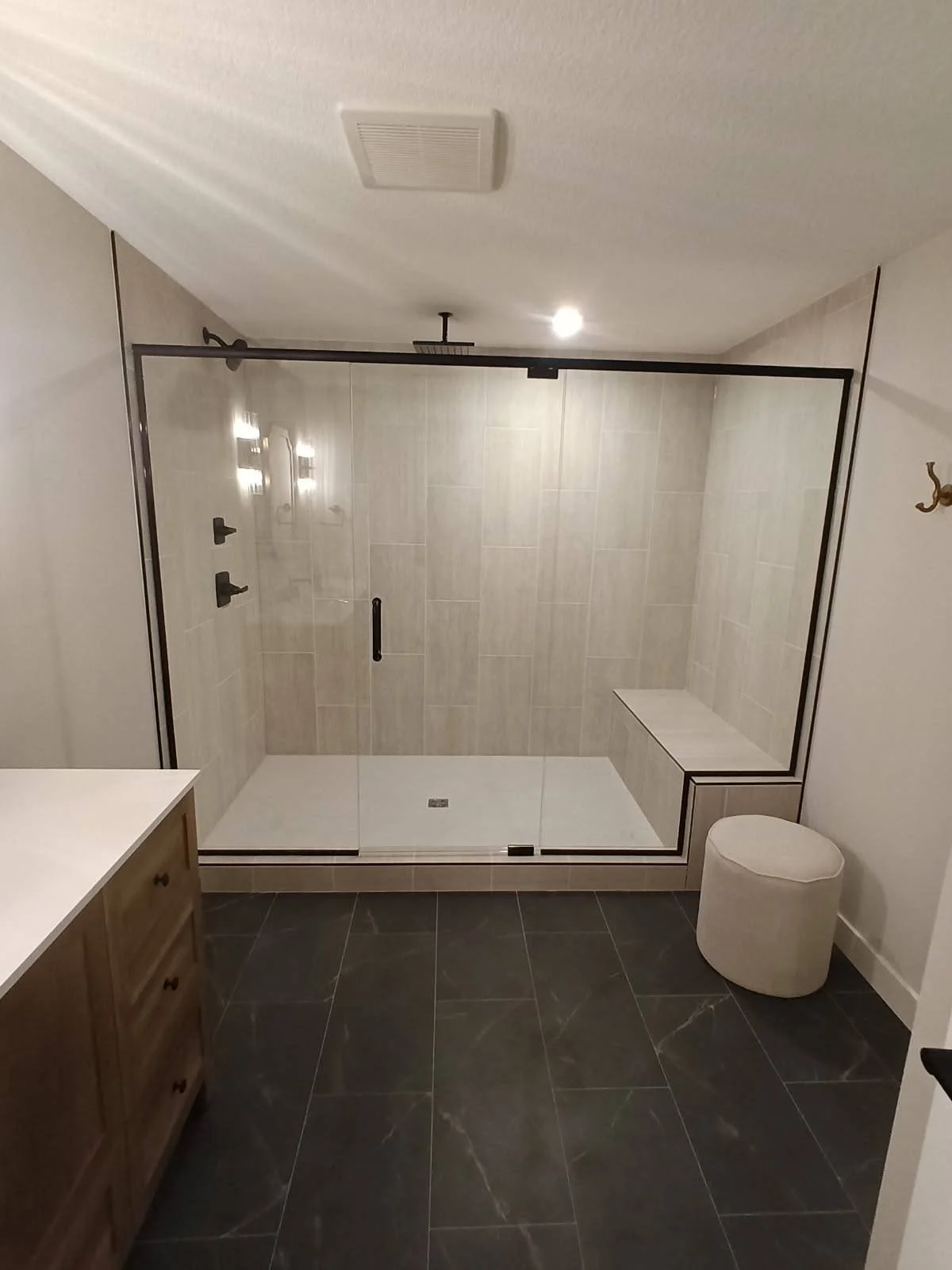 Modern bathroom with a large walk-in glass shower featuring beige tiles, a built-in bench, black hardware, and black-framed glass door. Dark marble floor, wooden vanity on the left, white round ottoman on the right, and a ceiling vent.