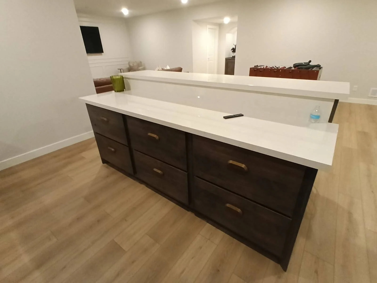 Kitchen island with white countertop and dark wooden drawers, a green glass, a black remote, and a water bottle on top.