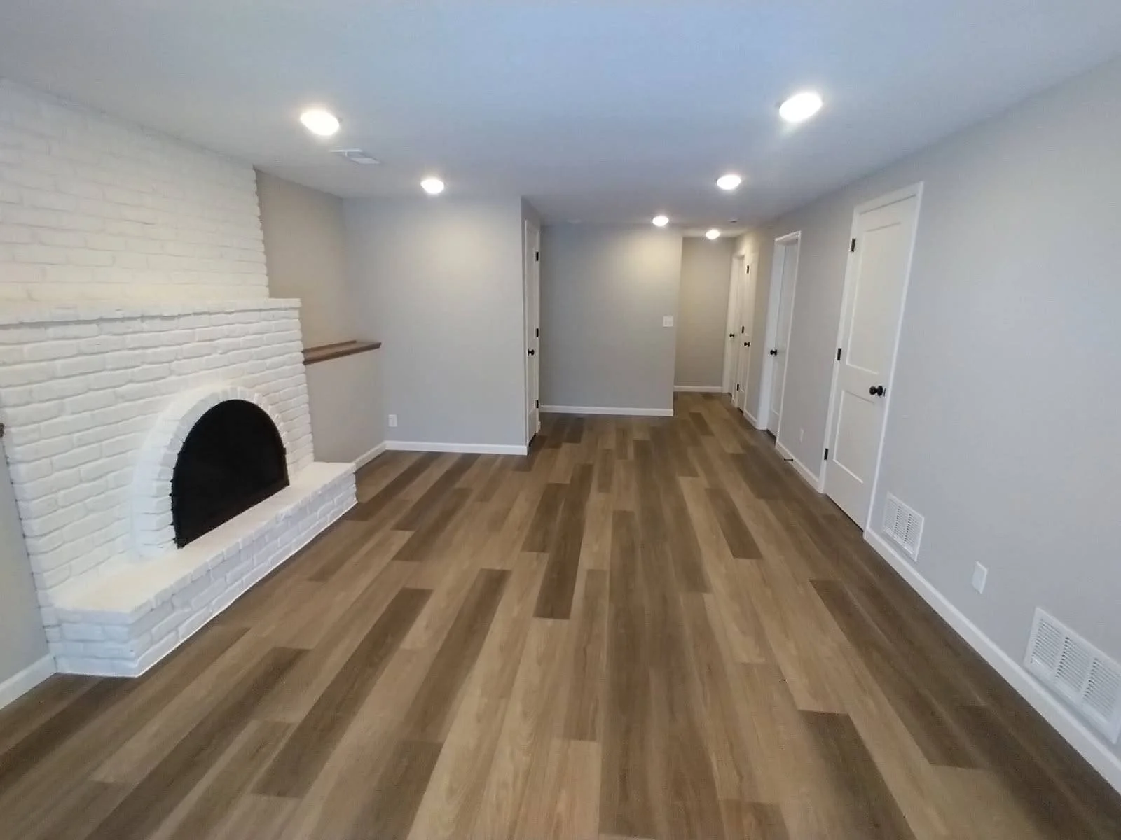 Empty living room with hardwood floors, white brick fireplace, gray walls, white doors, and ceiling lights.