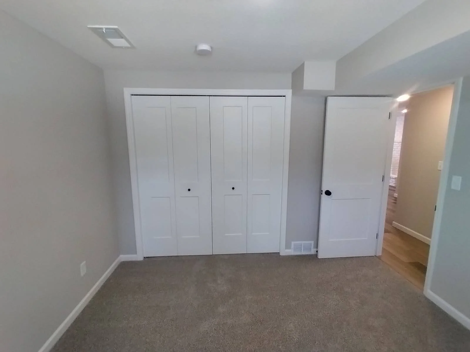 Empty beige-carpeted room with white double closet doors, an open white door leading to a hallway with wood flooring, and a ceiling vent.