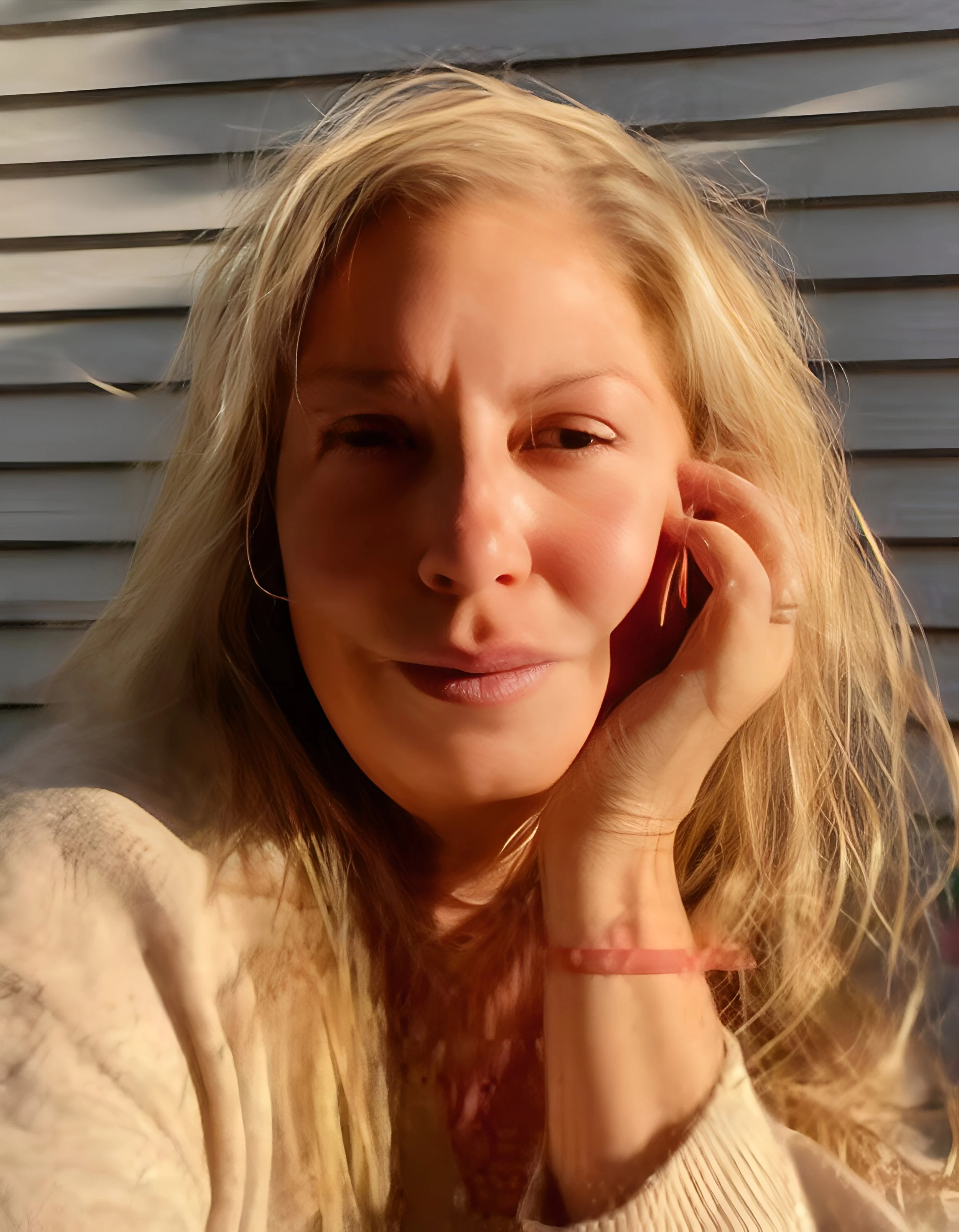 Close-up of a woman with blonde hair, outdoors, wearing a beige top, touching her hair with her right hand, and winking.