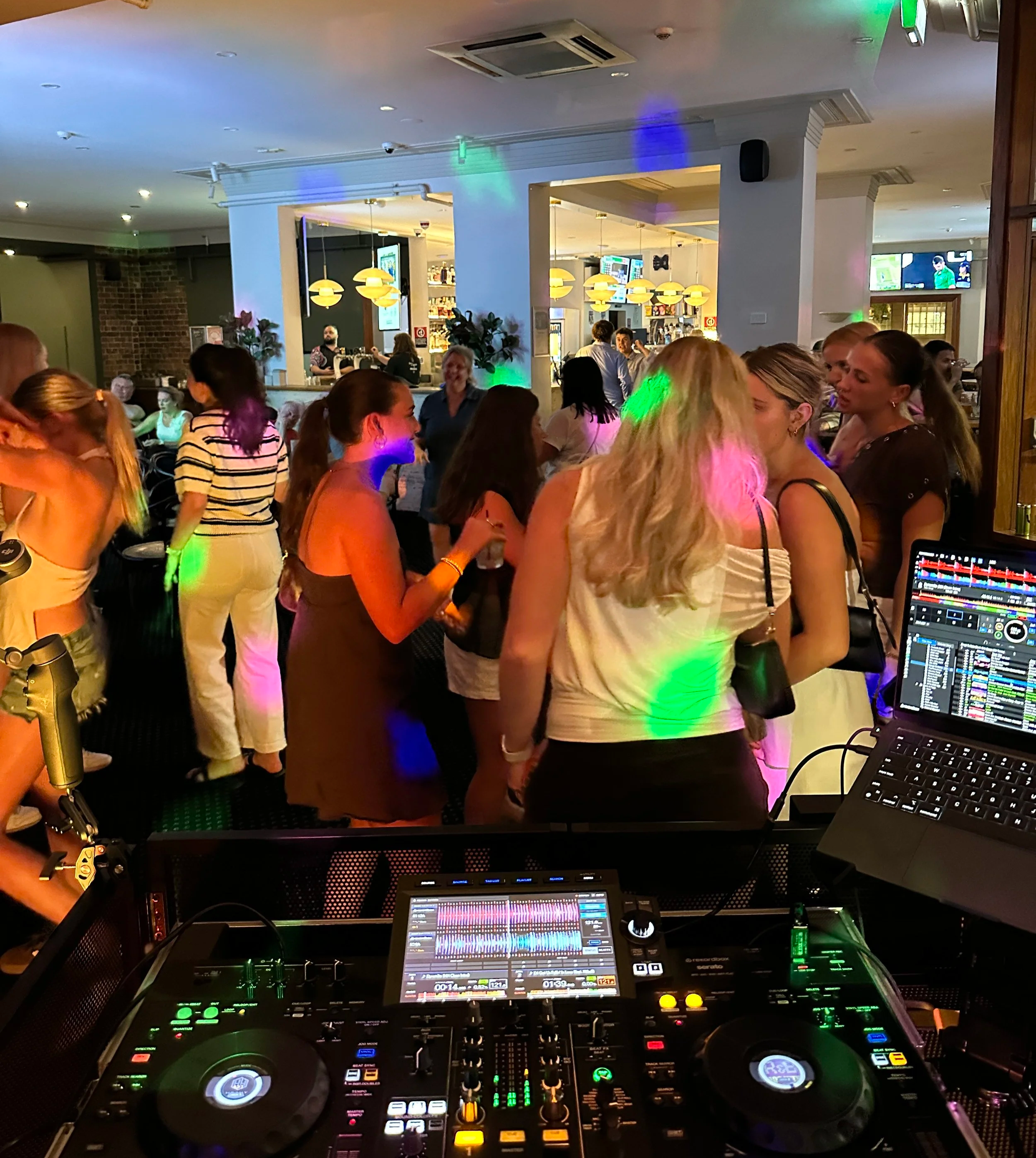 People dancing and socialising at a bar in Orange with DJ equipment in the foreground, colourful lights and a bar in the background.