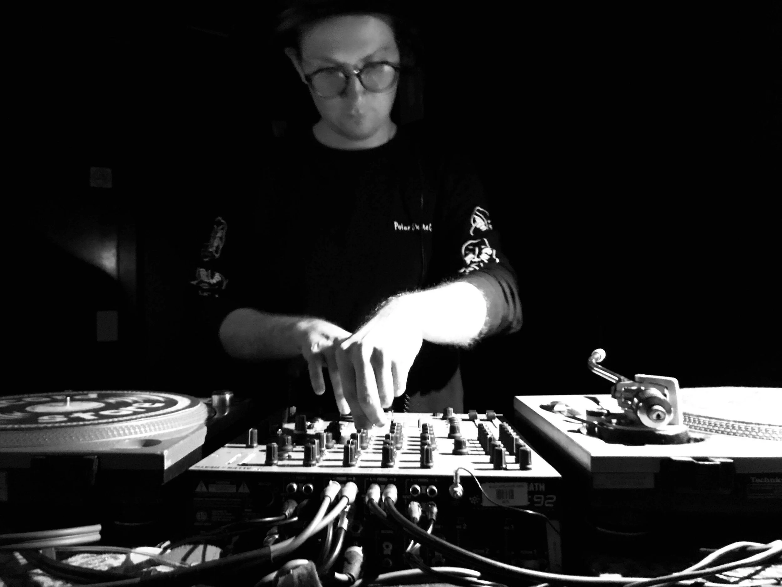 A DJ is mixing music at a darkened club, wearing glasses and a black shirt, with turntables and audio equipment in front of him.