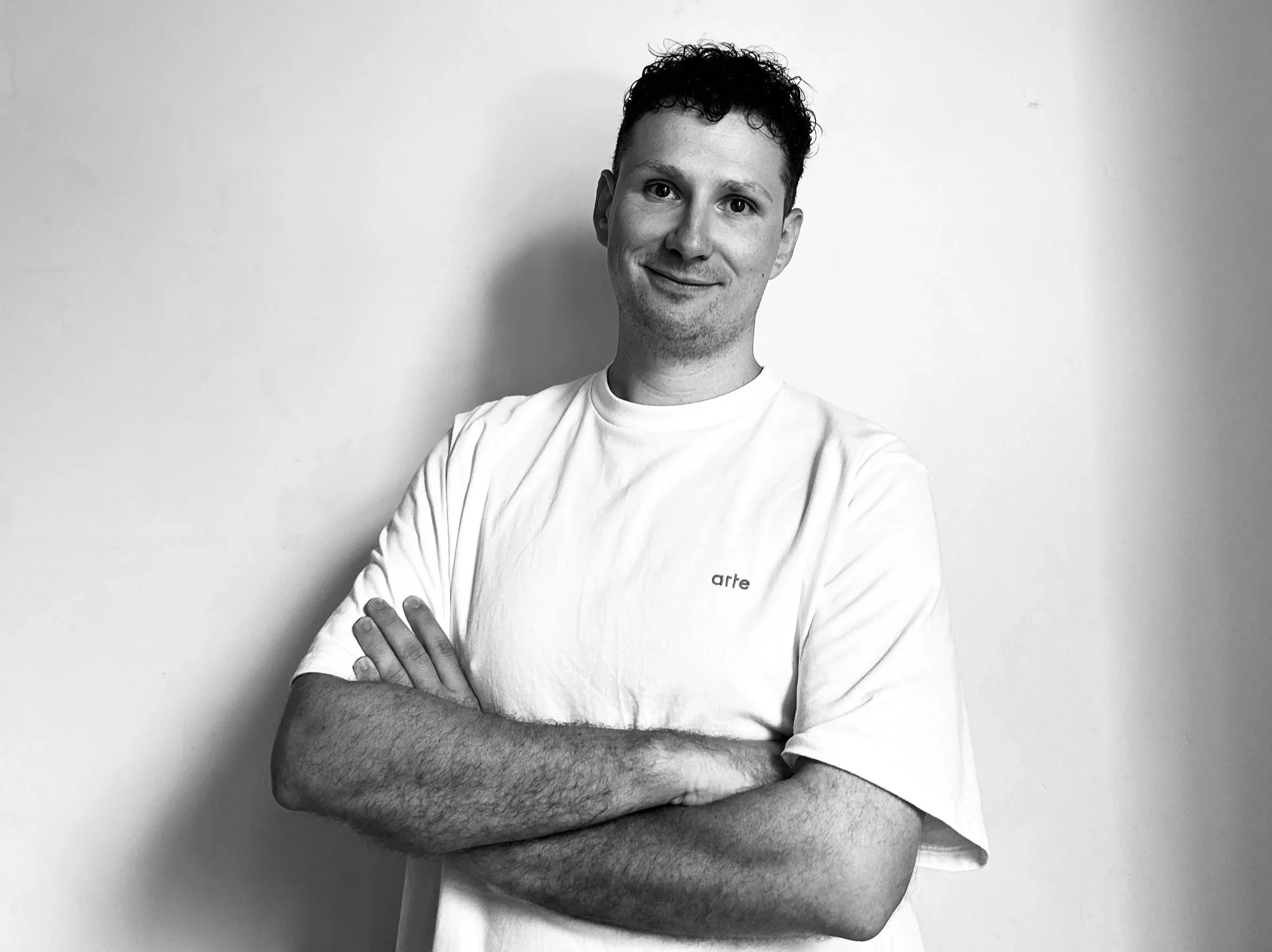 Black and white portrait of a young man smiling, arms crossed, wearing a white t-shirt with small text 'arte' on it, standing against a plain wall.