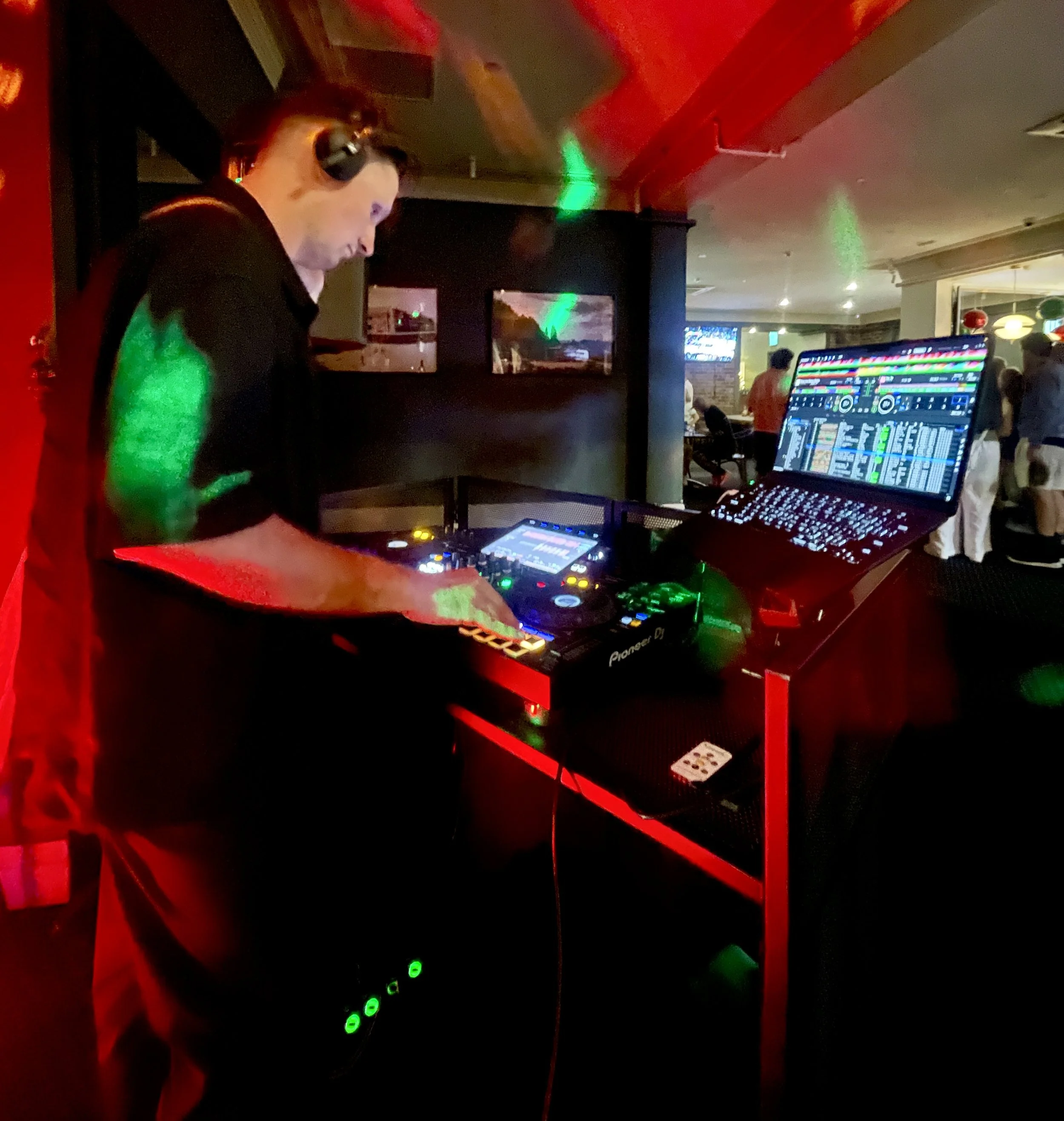 A DJ wearing headphones performing at a nightclub with colorful lighting, using a Pioneer DJ controller and a laptop with a digital DJ software interface.