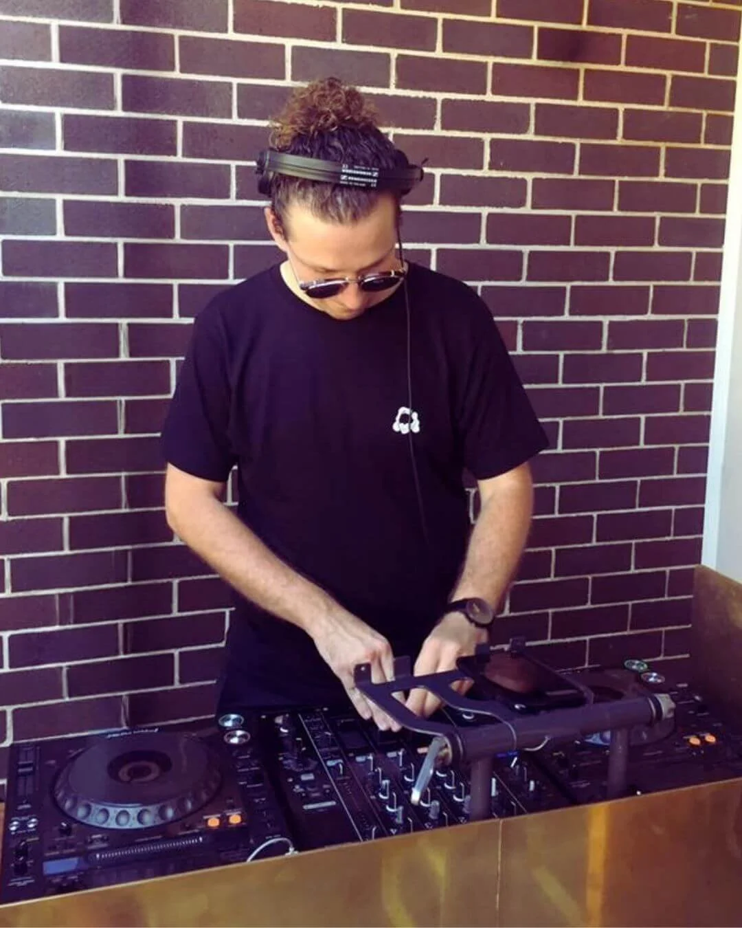 A DJ wearing sunglasses, headphones, and a black t-shirt is mixing music with a DJ controller against a brick wall.