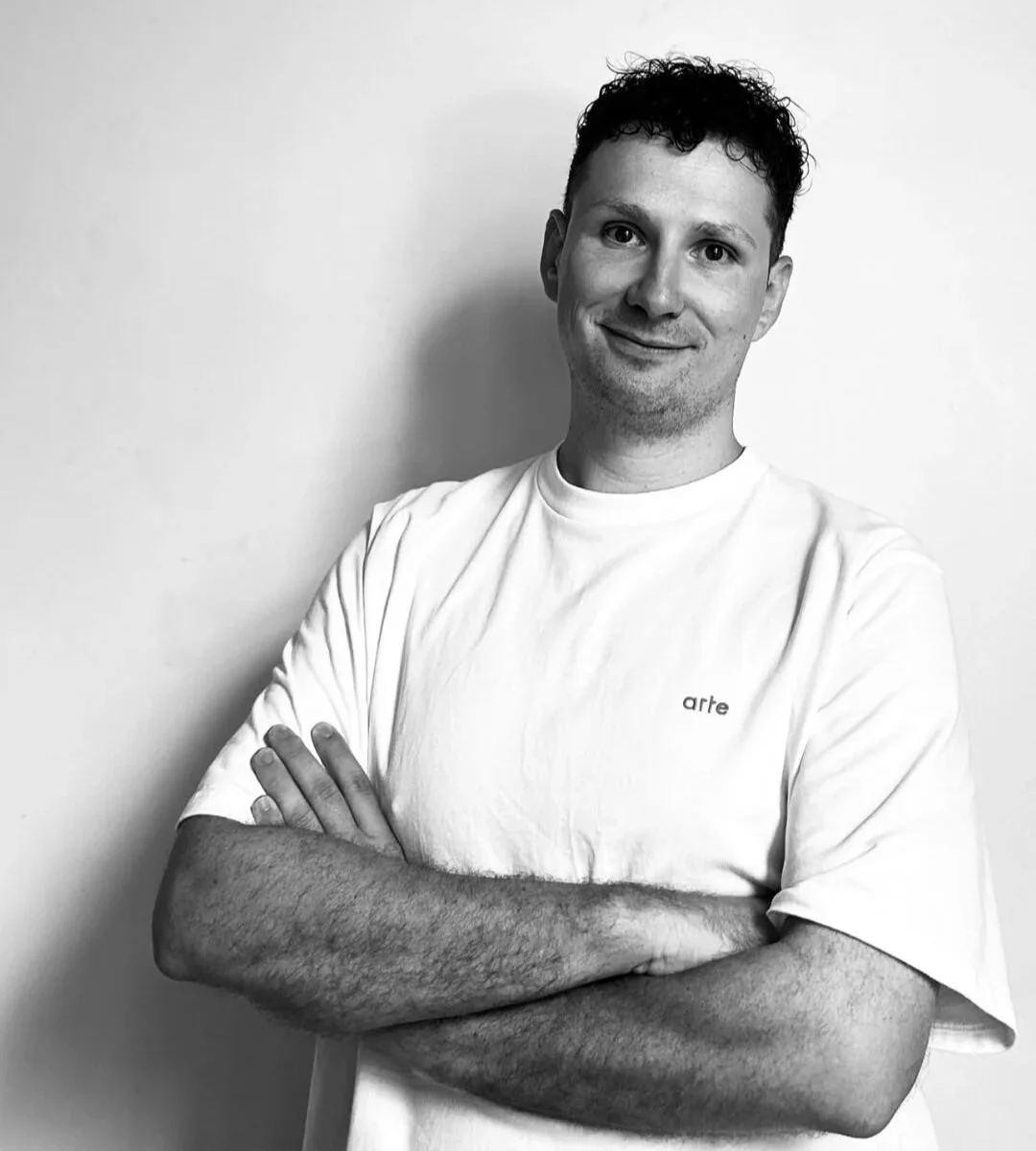 Black-and-white photo of a young man with short dark curly hair smiling and crossing his arms, wearing a white T-shirt with the word 'arte' on it, standing against a plain white wall.