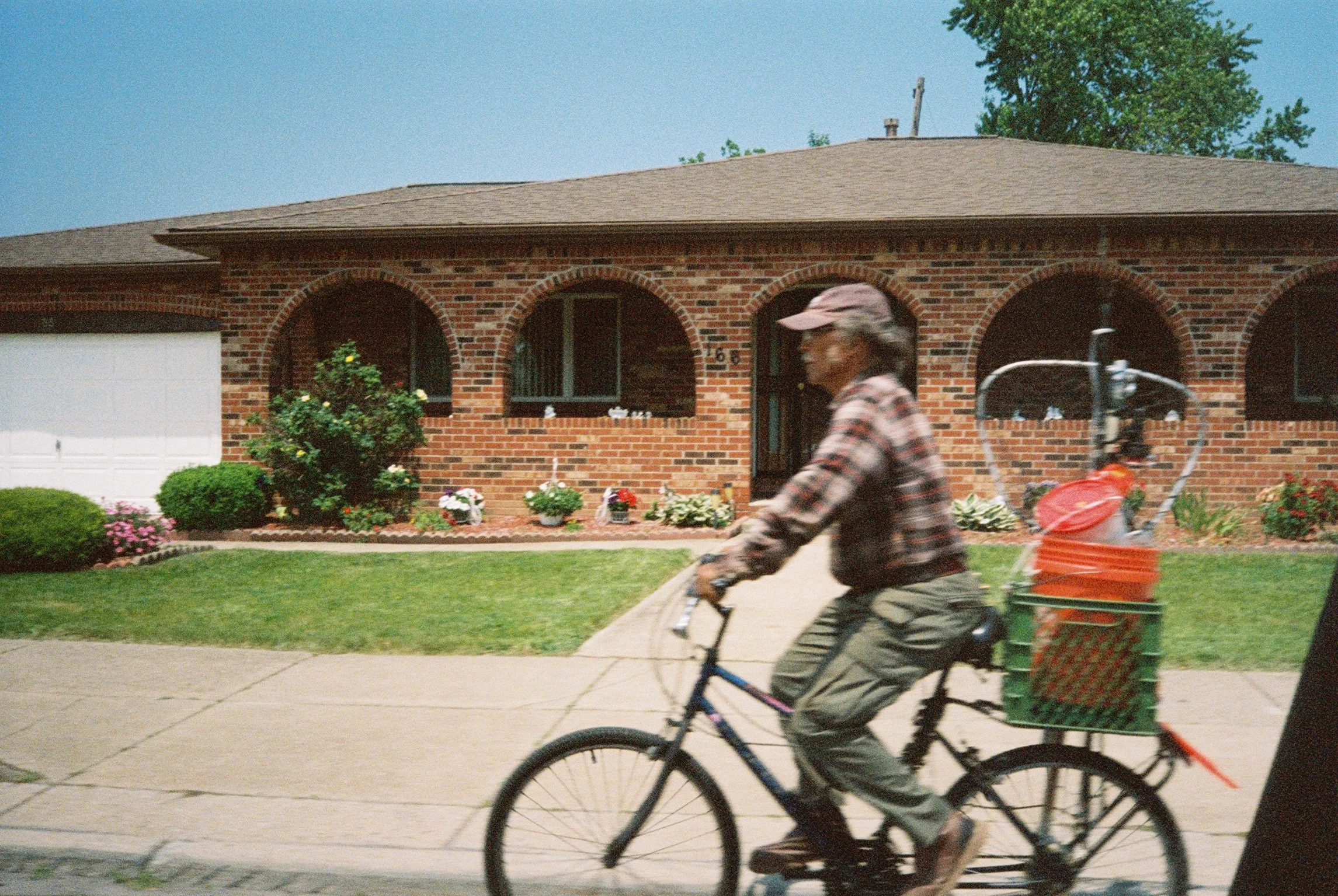 A man riding a bicycle with a large basket attached to the back, filled with buckets and a container, in front of a red brick house with arched windows and a garden.
