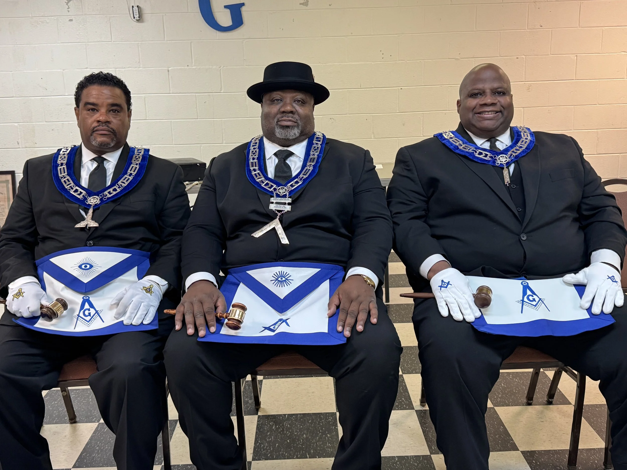 Three men in black suits with Masonic regalia, sitting side by side in a room with tiled black and white floor, each holding a small gavel and wearing white gloves, Masonic aprons, and blue collars with silver chains.