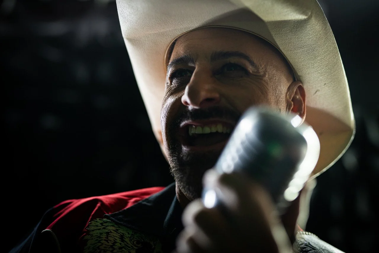 A man wearing a cowboy hat and a red shirt, laughing while holding a microphone close to his mouth.