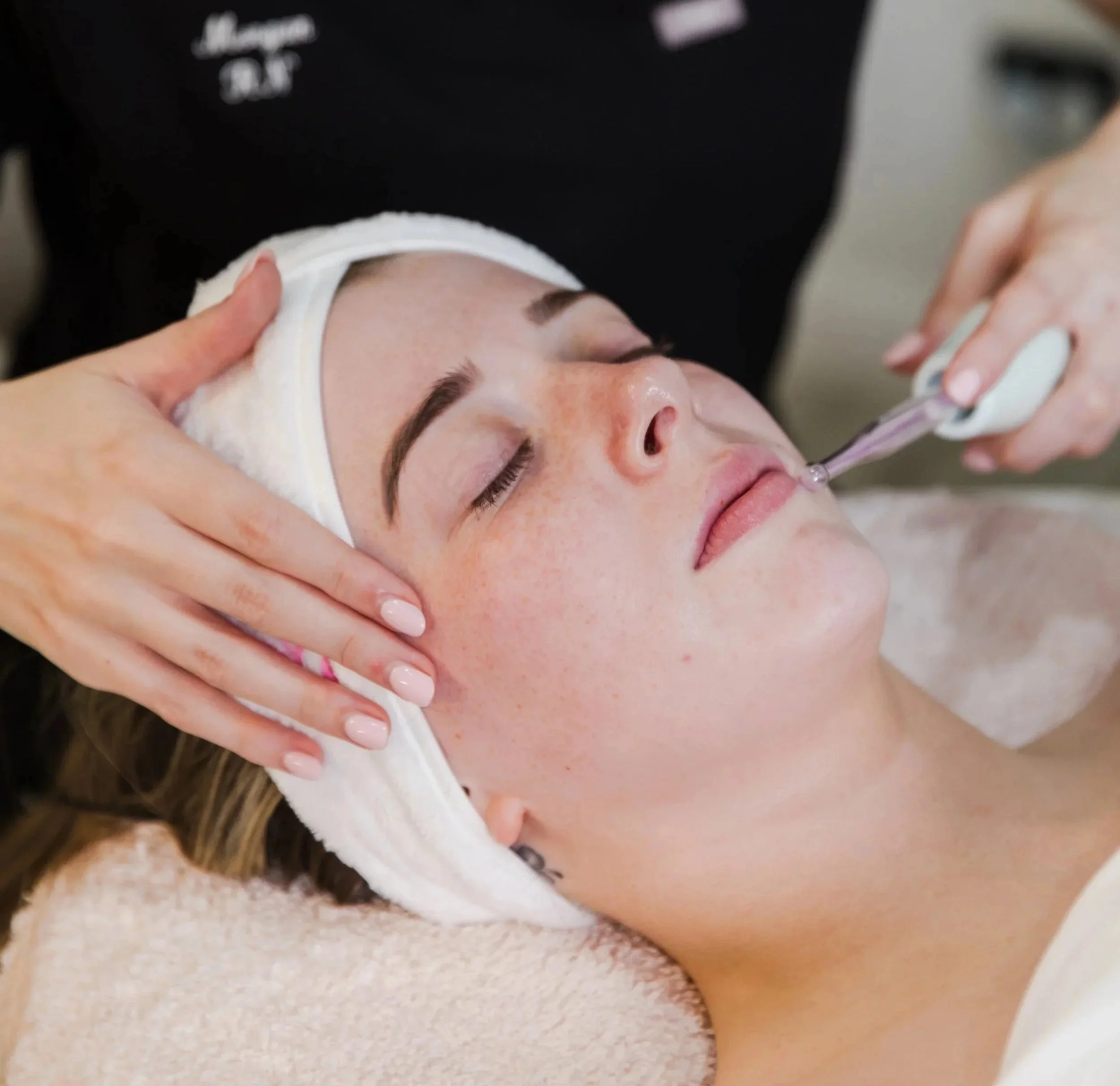 A woman receiving a facial treatment, lying down with her eyes closed, a towel wrapped around her head, as a skincare professional doing a high frequency treatment