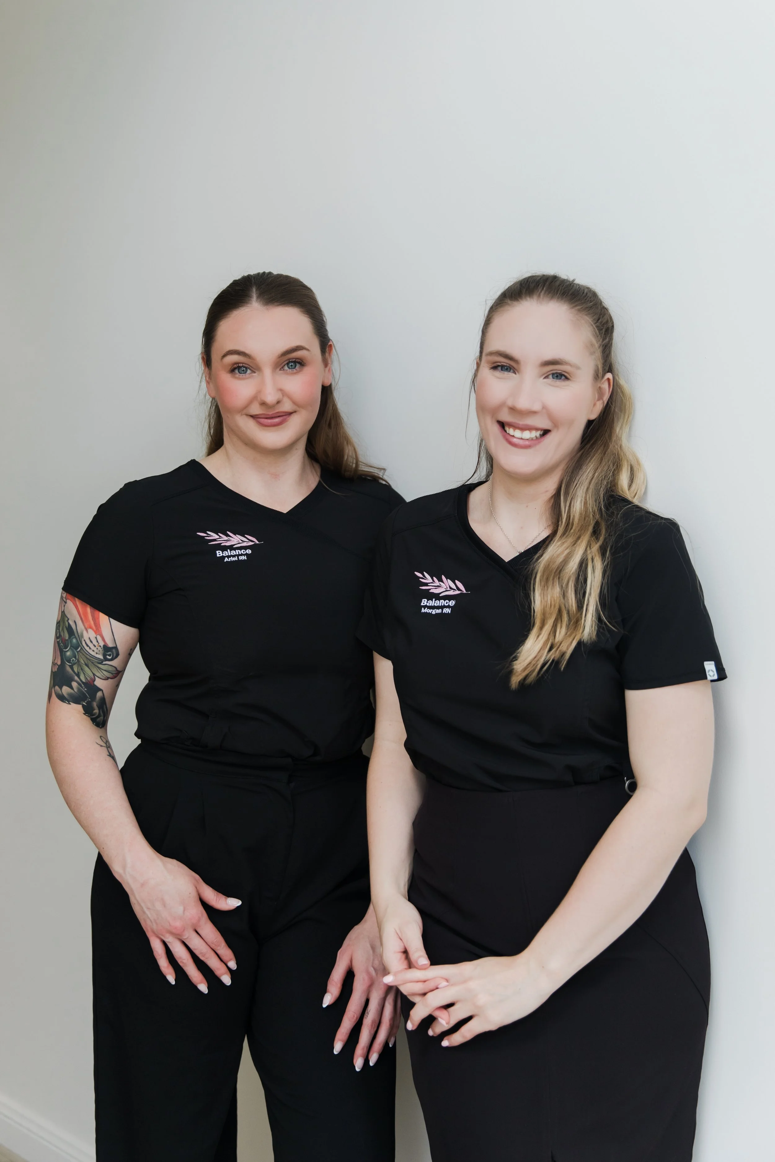Two women in black scrubs standing side by side against a white wall, smiling at the camera. The woman on the left has dark hair and a colorful tattoo sleeve on her left arm. The woman on the right has blonde hair.