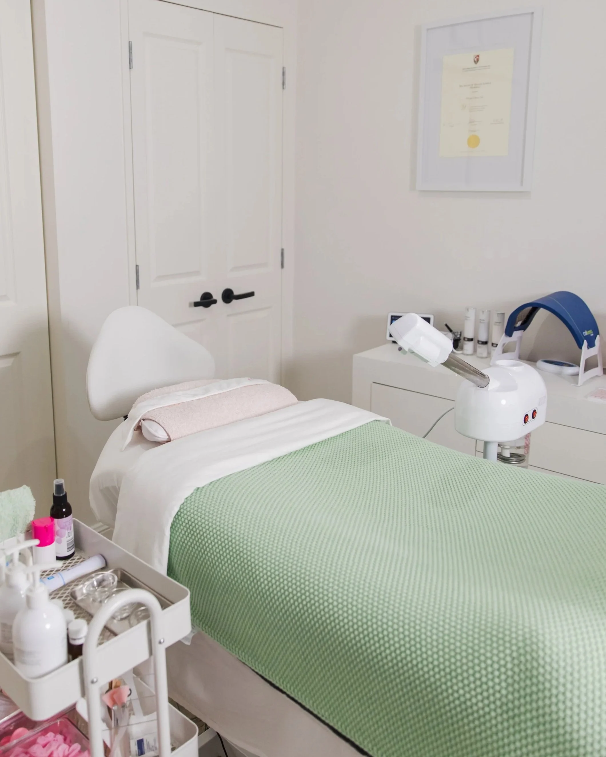 A medical or spa treatment room featuring a single bed with white sheets, a pink towel, and a green blanket, along with medical supplies on a nearby cart and equipment on a white counter.