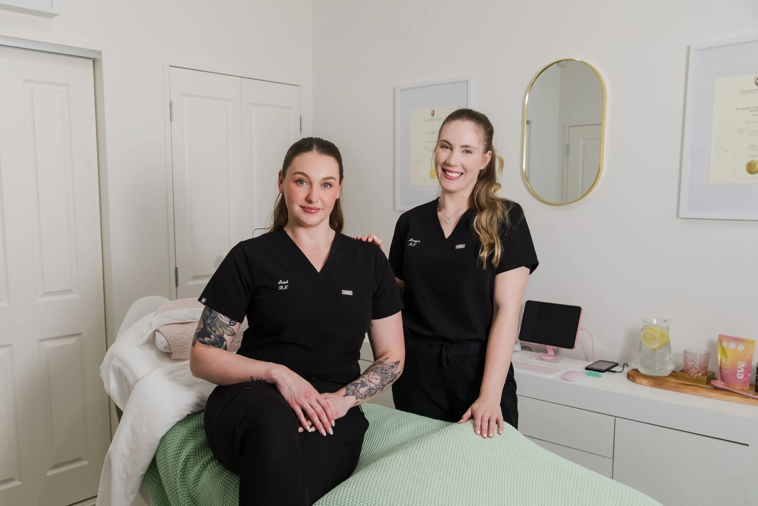 Two smiling women in black scrubs posing inside a medical or skincare clinic room. One woman is seated on a bed with a green blanket, and the other is standing beside her. The wall behind them has framed certificates and a mirror.