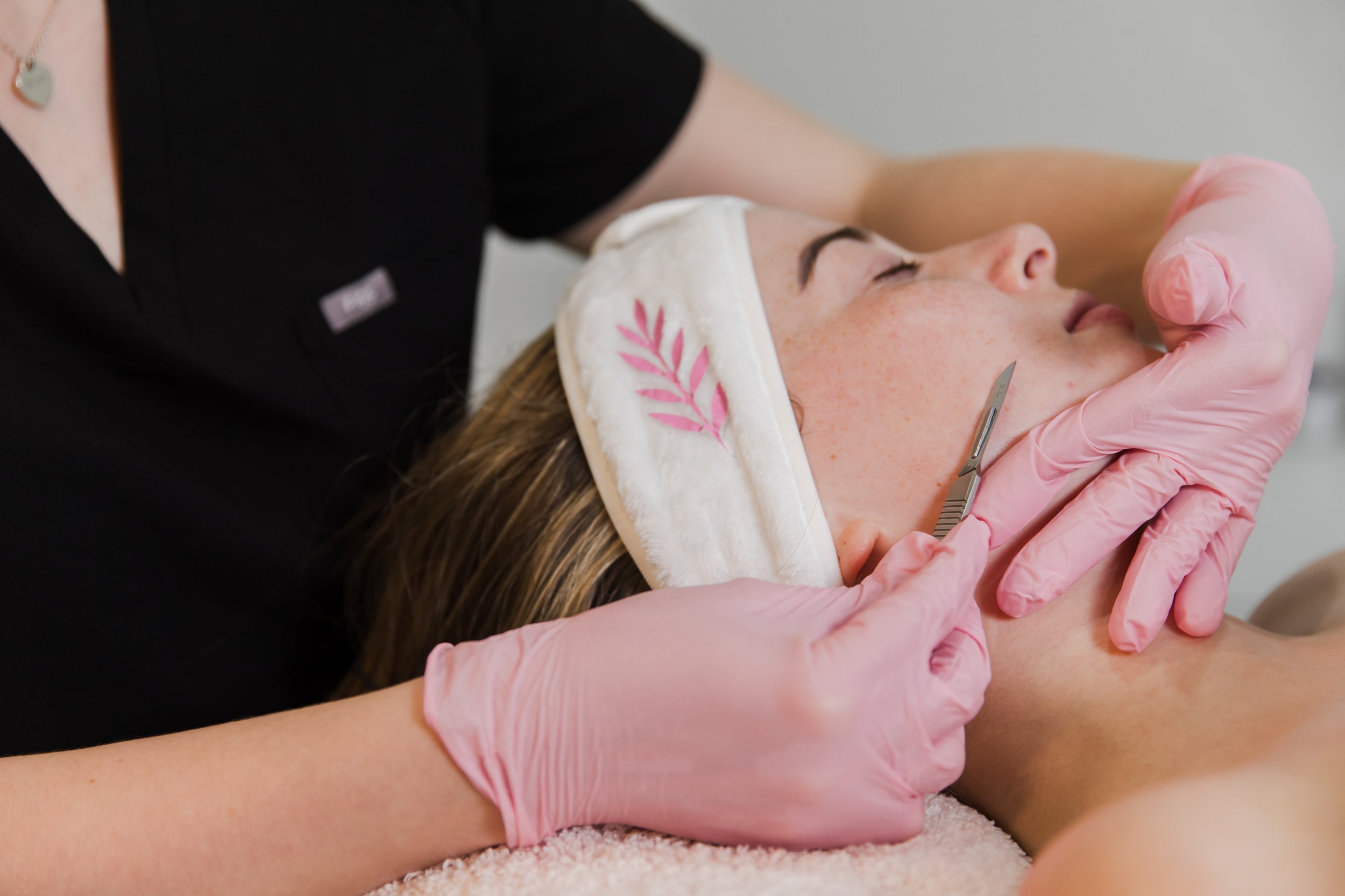 A woman receiving a facial treatment from an esthetician in a spa setting. The woman is lying down with her eyes closed, wearing a headband, while the esthetician uses scalpel to provide Dermablading.