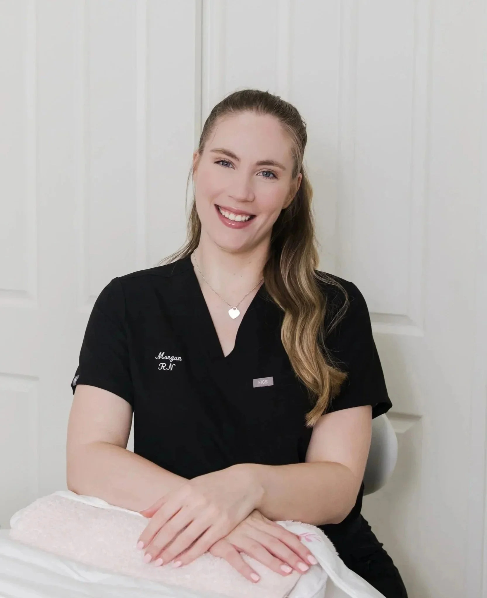 A smiling woman with long wavy hair in a black nursing uniform sitting at a table with her hands resting on a folded towel.