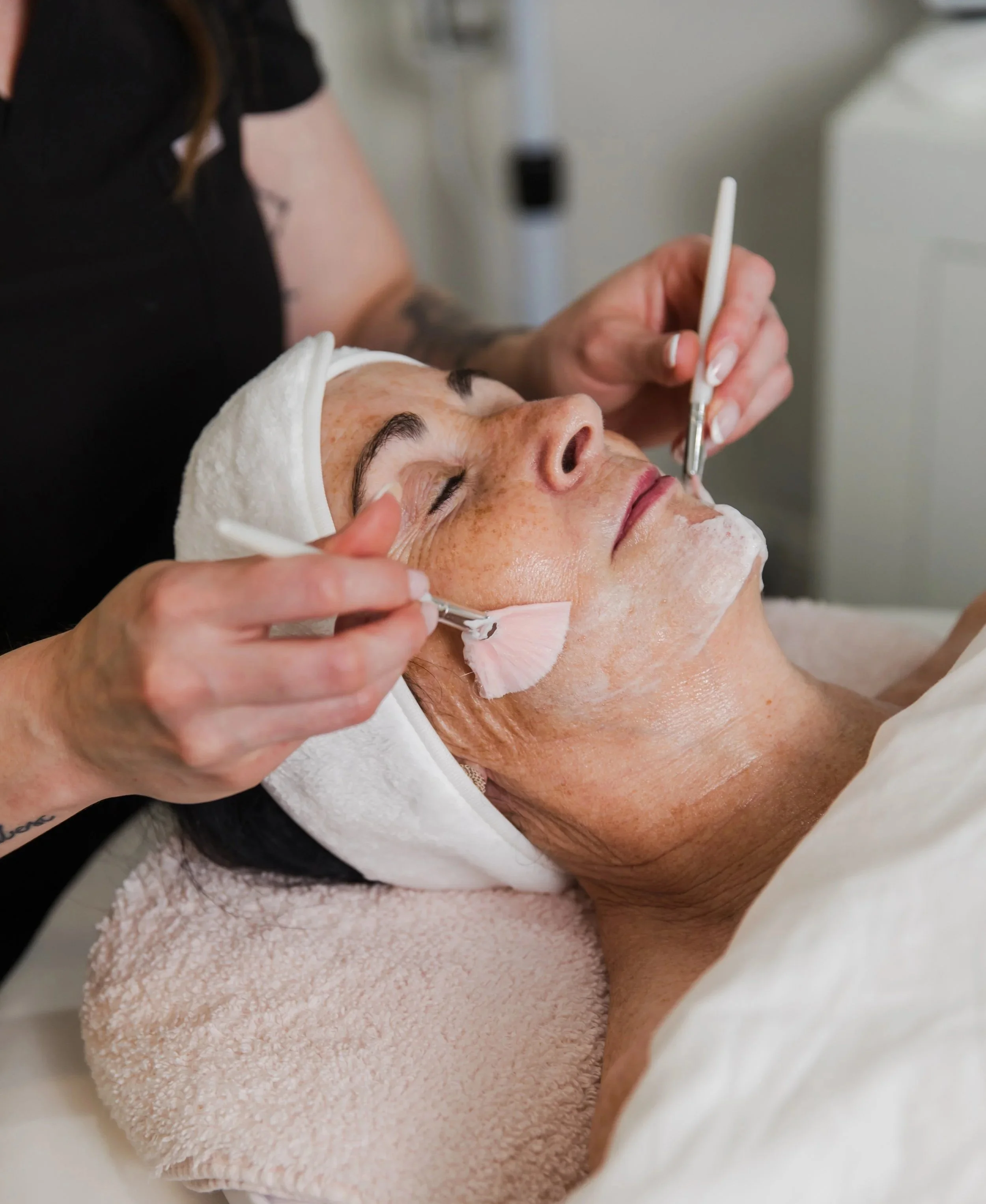 Woman receiving a facial treatment, lying down with eyes closed, a towel wrapped around her head, as a skincare professional applies skincare products with brushes.