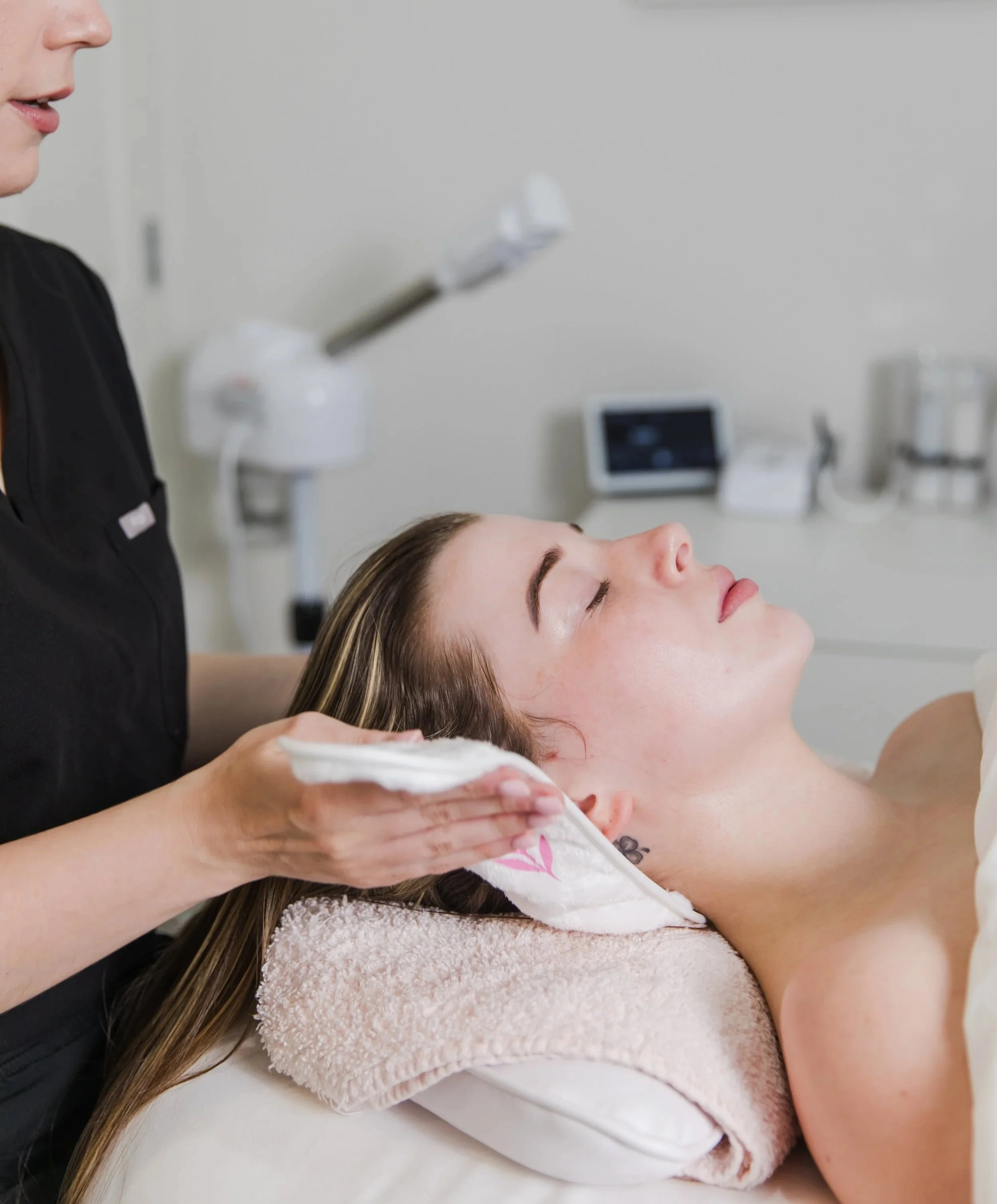 A woman receiving a facial treatment at a spa or skincare clinic, lying on a treatment bed with an aesthetician wiping her face with a cloth.