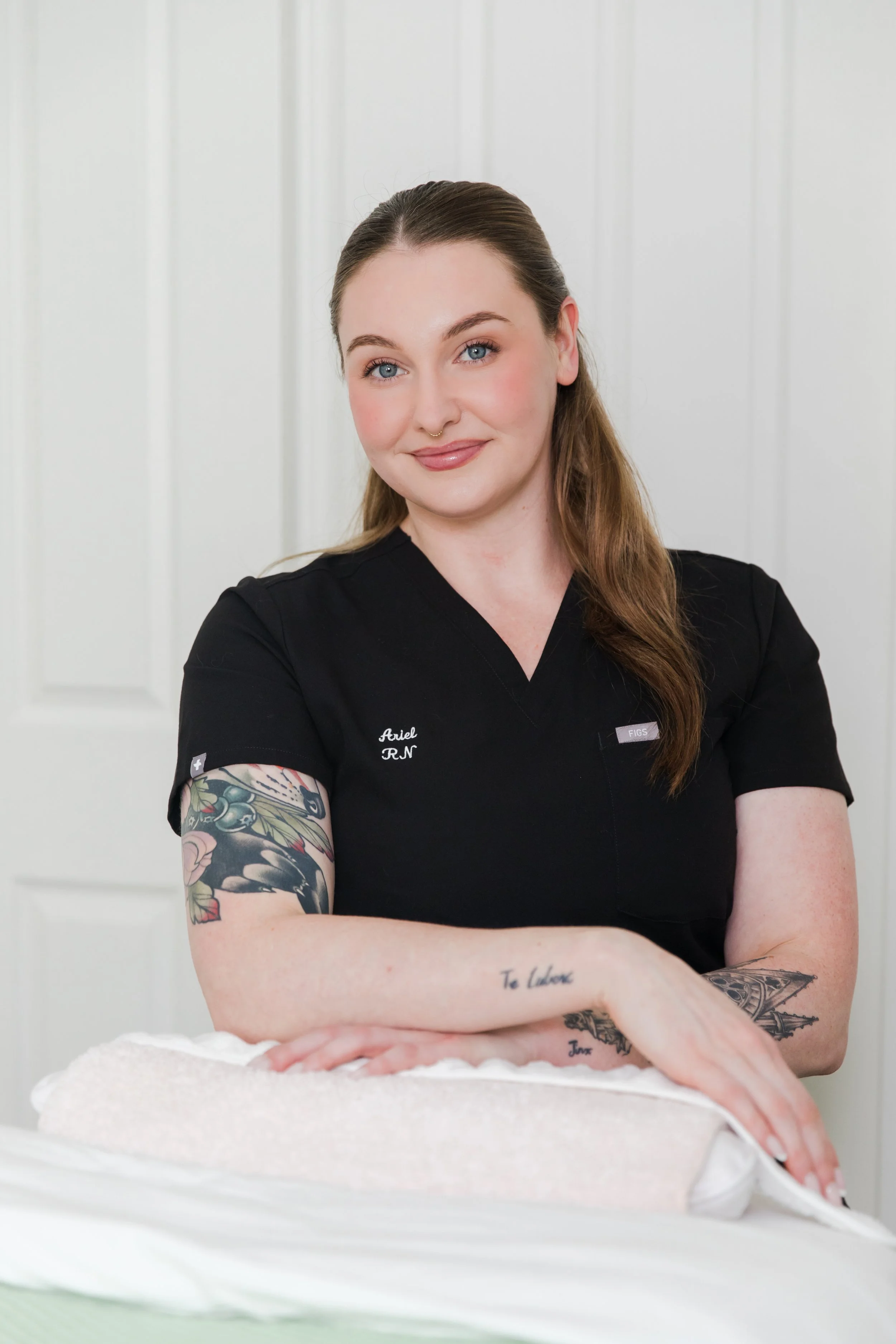 A woman with long hair, tattoos, and a black medical uniform, smiling and leaning on a bed or table, in a bright room.