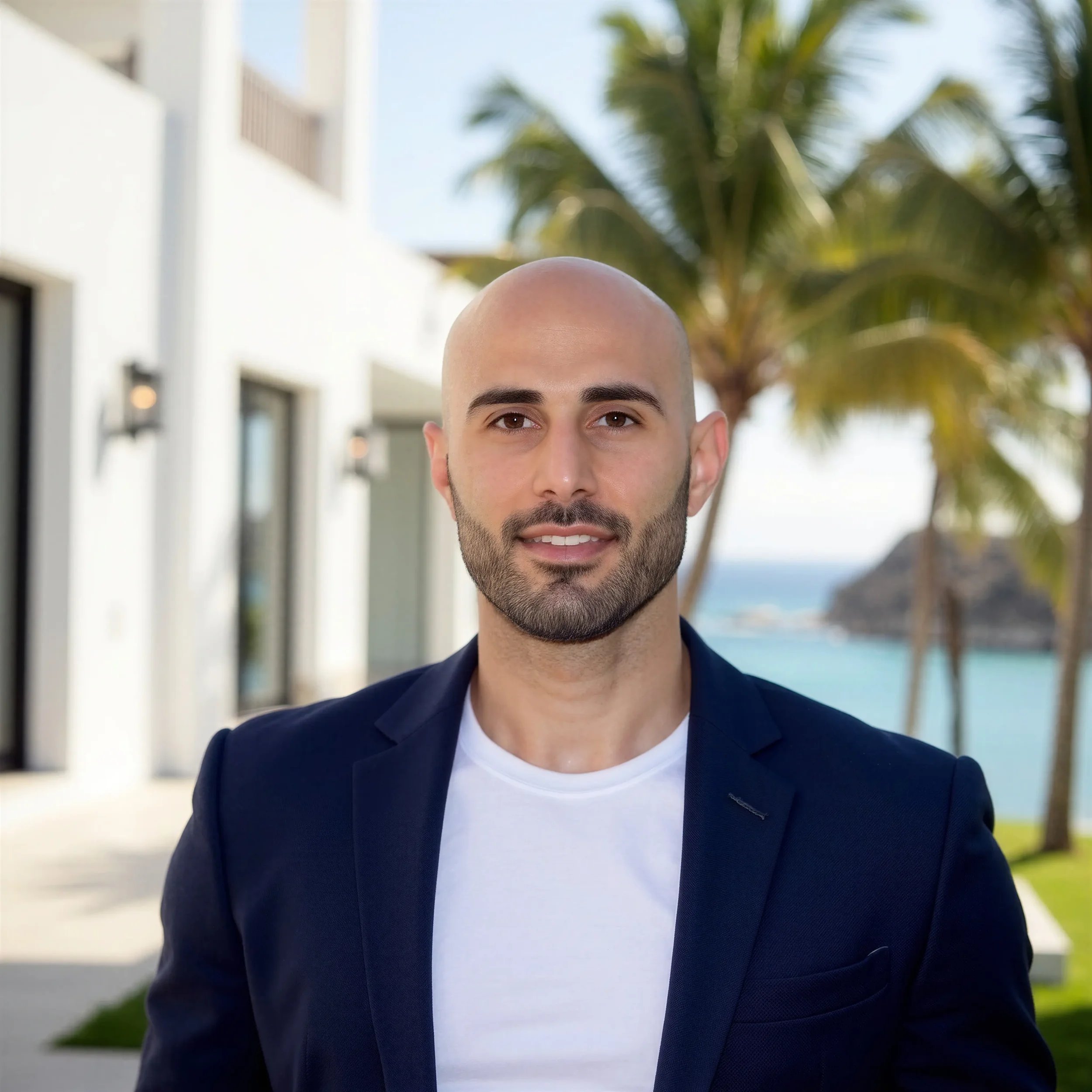 Close-up of a man in a navy blazer and white t-shirt standing outdoors near palm trees, with a beach and ocean in the background.