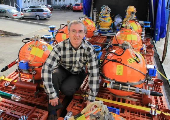 A man crouches next to several orange and yellow underwater diving chambers and equipment on a red pallet at an outdoor event.