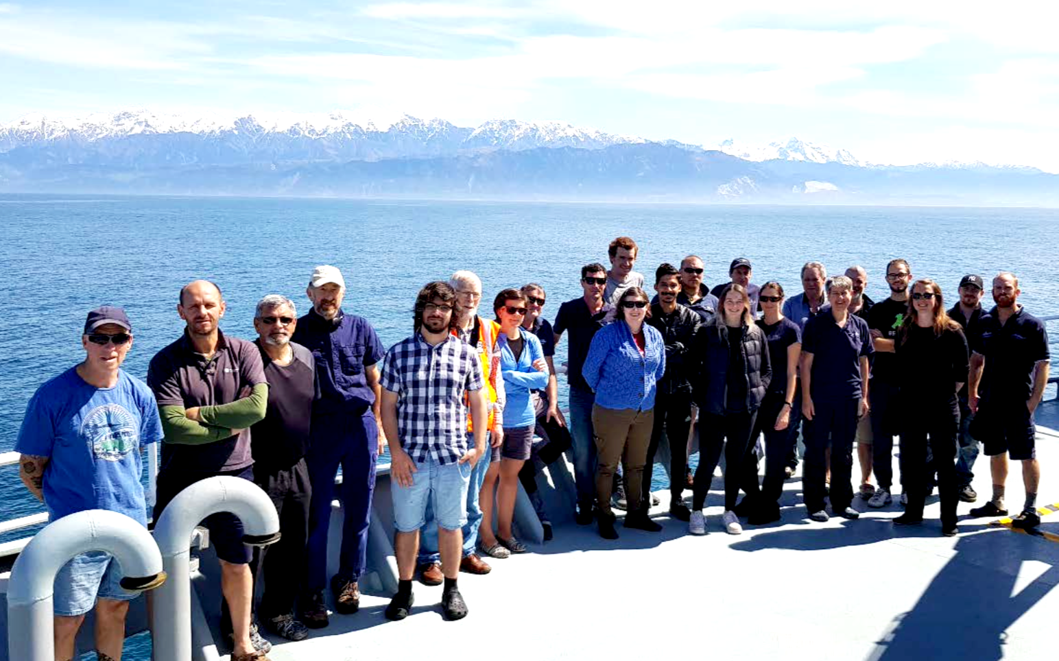 Group of people standing on a ship's deck with a body of water and snow-capped mountains in the background.