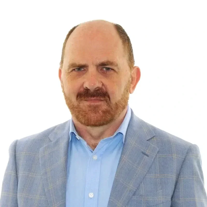 A middle-aged man with a reddish beard and receding hairline, wearing a light blue dress shirt and a light gray blazer, standing against a plain white background.