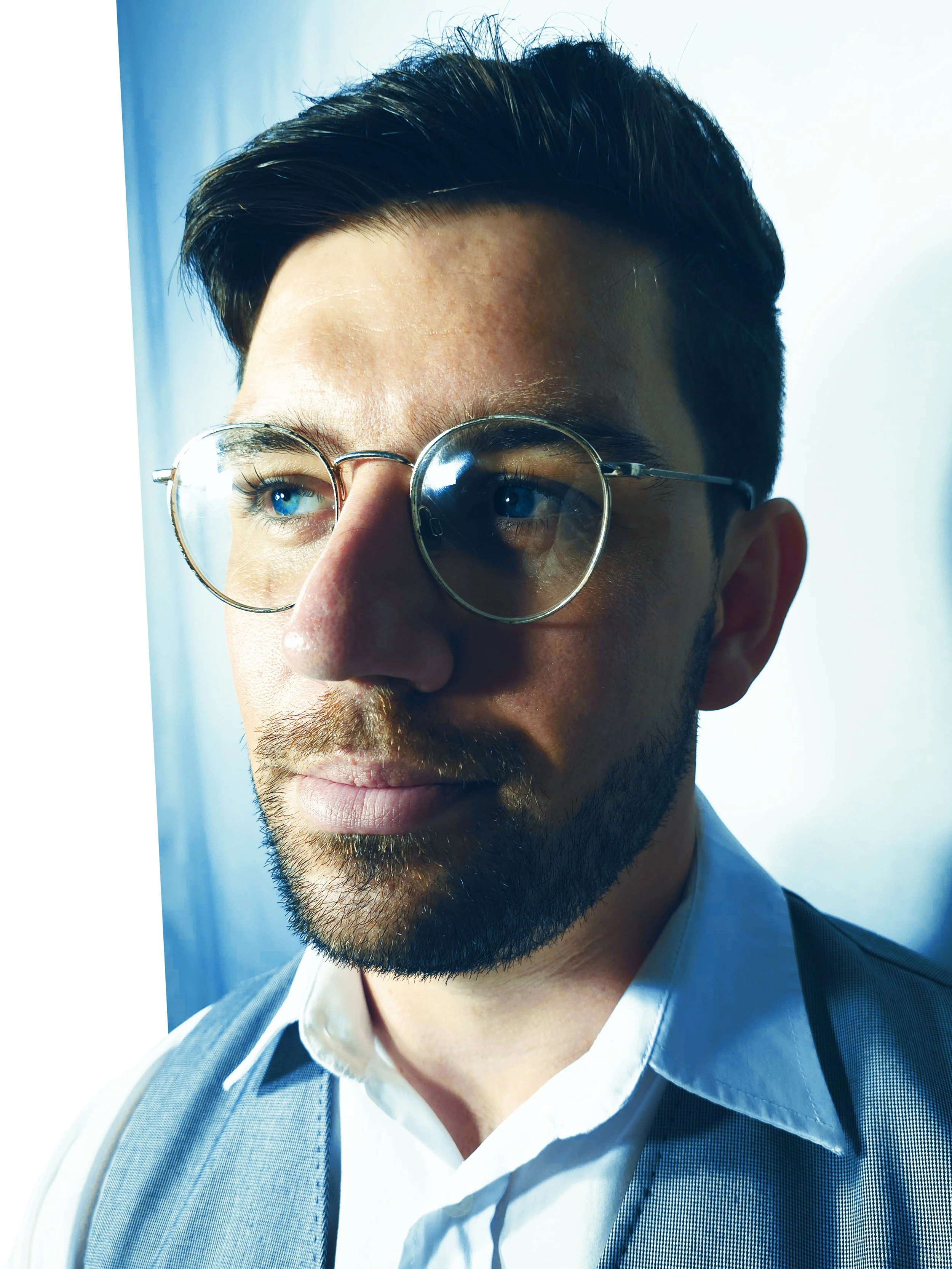 Close-up of a man with glasses, brown hair, and a beard, looking out a window with natural light illuminating his face.