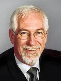 Portrait of an elderly man with white hair and glasses, smiling, wearing a suit and tie.