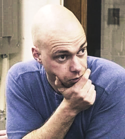 A bald man with a pensive expression resting his chin on his hand, wearing a blue shirt.