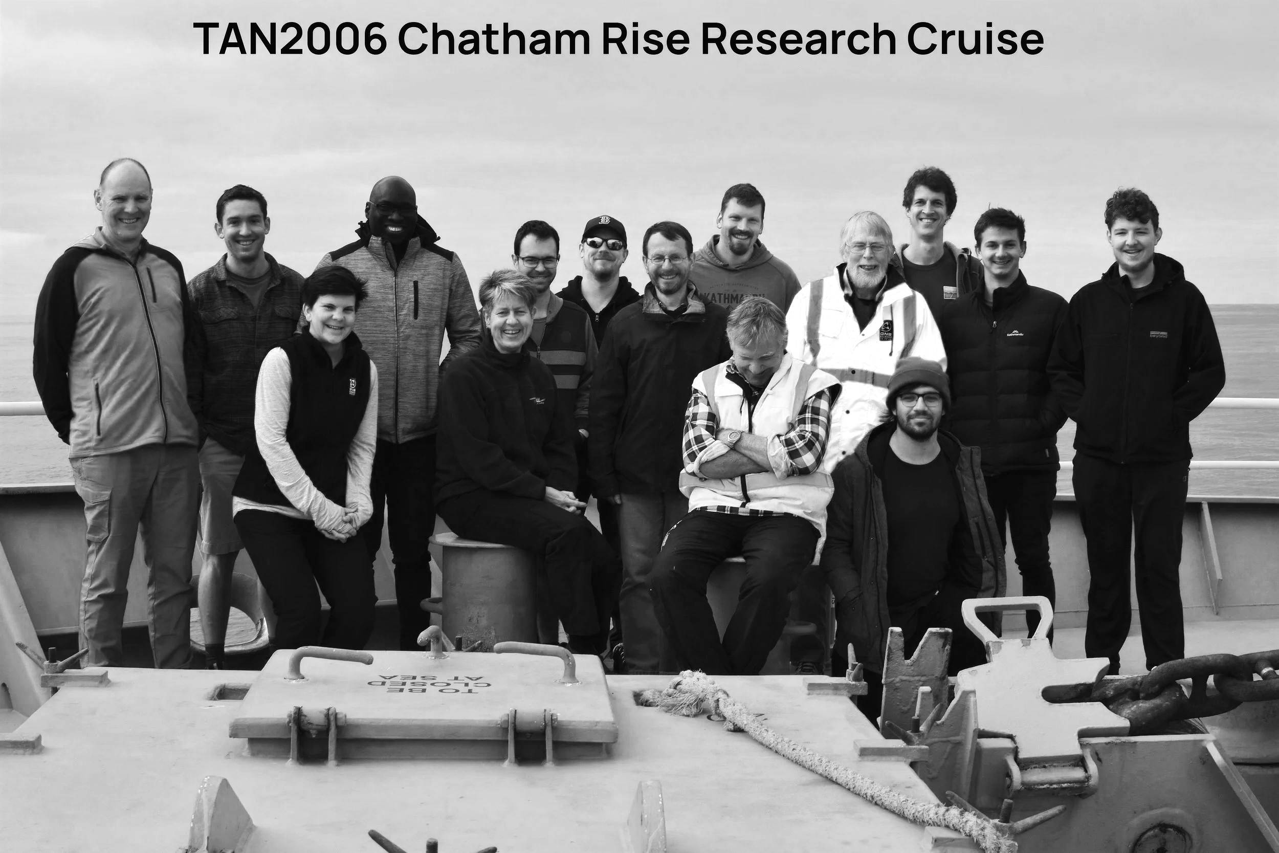 A group of 14 people on a research cruise ship deck, with some seated and others standing, posing for a photo during the TAN2006 Chatham Rise Research Cruise, with the ocean in the background.