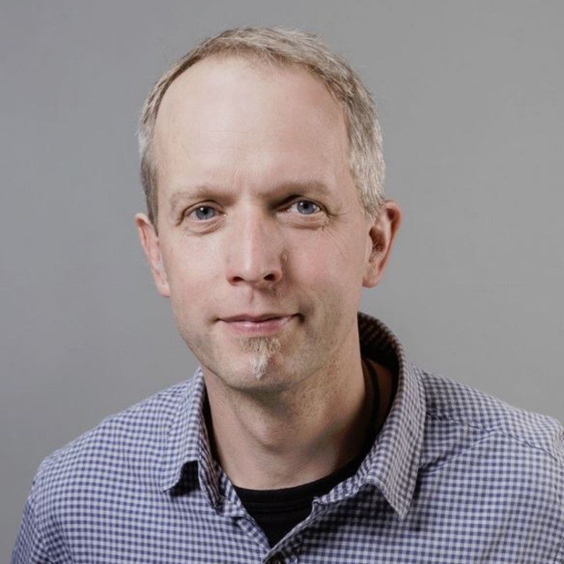 Portrait of a middle-aged man with short, light-colored hair, blue eyes, and a small goatee, wearing a checkered shirt against a plain gray background.