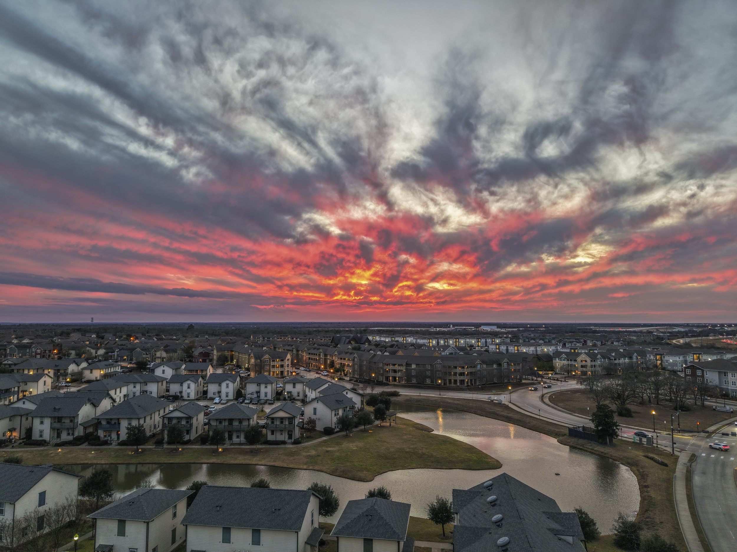 Sunset over a suburban neighborhood with houses, roads, and a pond, colorful sky with clouds in shades of pink, orange, and gray in College Station, Texas.