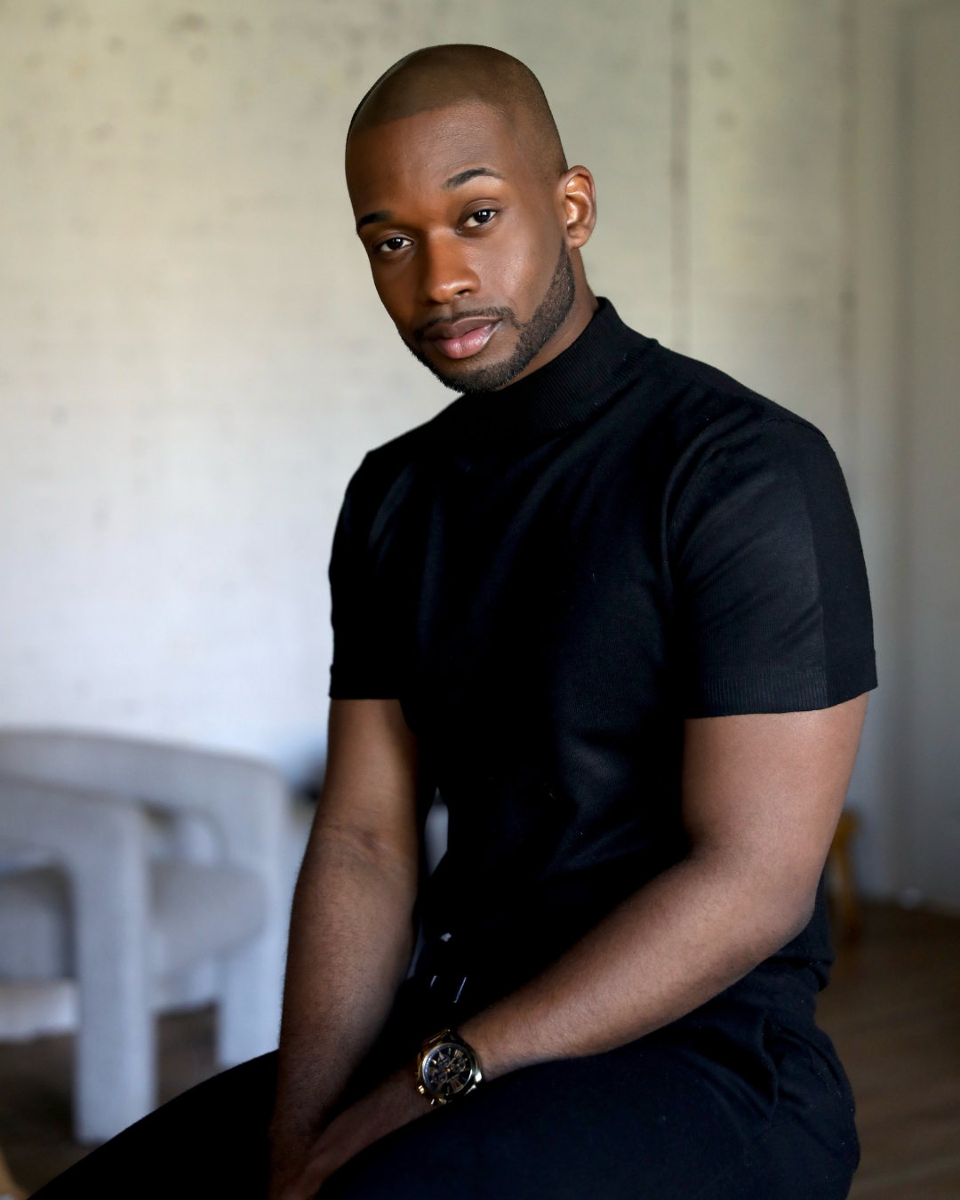 A young man with a shaved head and beard wearing a black T-shirt, sitting indoors with a neutral background.