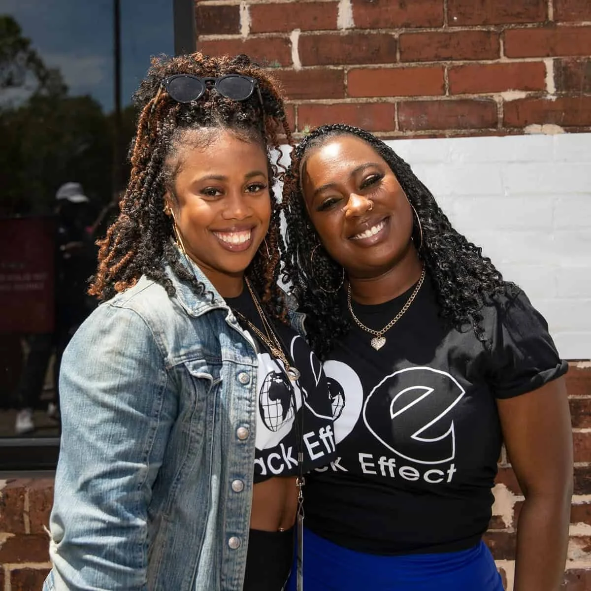 Two women smiling and standing close together outside, against a brick wall. The woman on the left has curly hair, sunglasses on her head, a denim jacket, and jewelry. The woman on the right has curly hair, hoop earrings, a necklace, and is wearing a