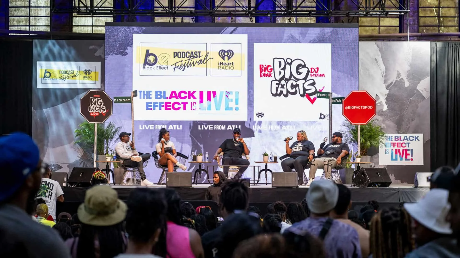 Panel discussion at a live event with five speakers seated on stage, large screen behind them displays promotional graphics for a podcast festival and Black Effect Podcast Network, audience members in foreground.