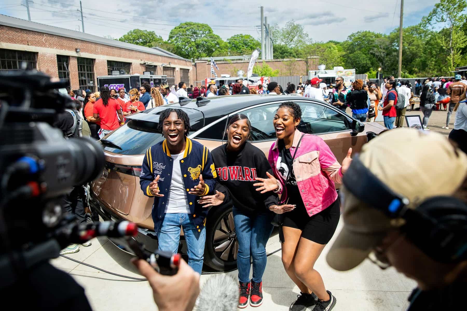Three women smiling and posing in front of a copper-colored car at an outdoor event, with a crowd of people in the background.