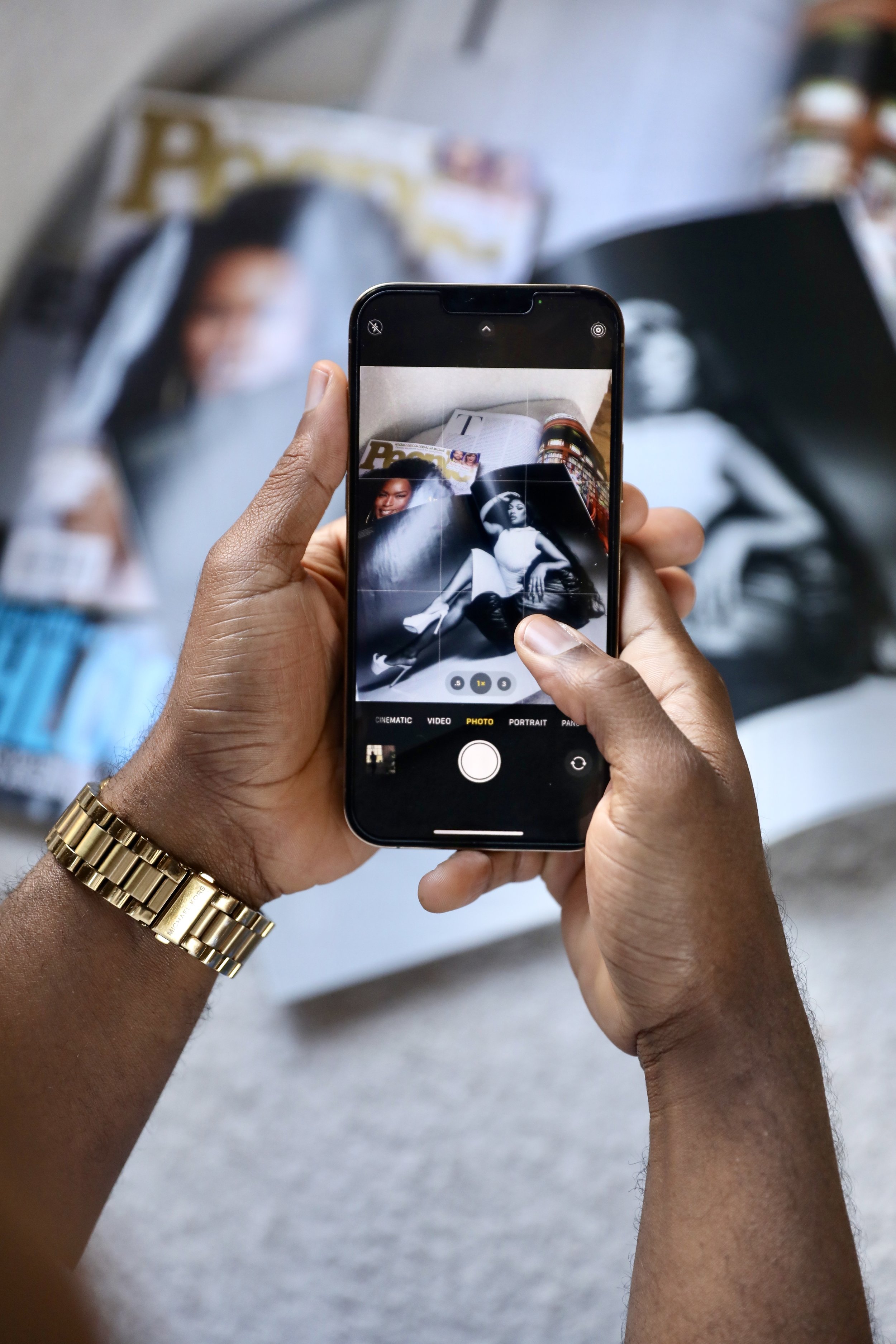 A person with a gold bracelet on their wrist holding a smartphone, taking a photo of fashion magazines and photographs on a table.
