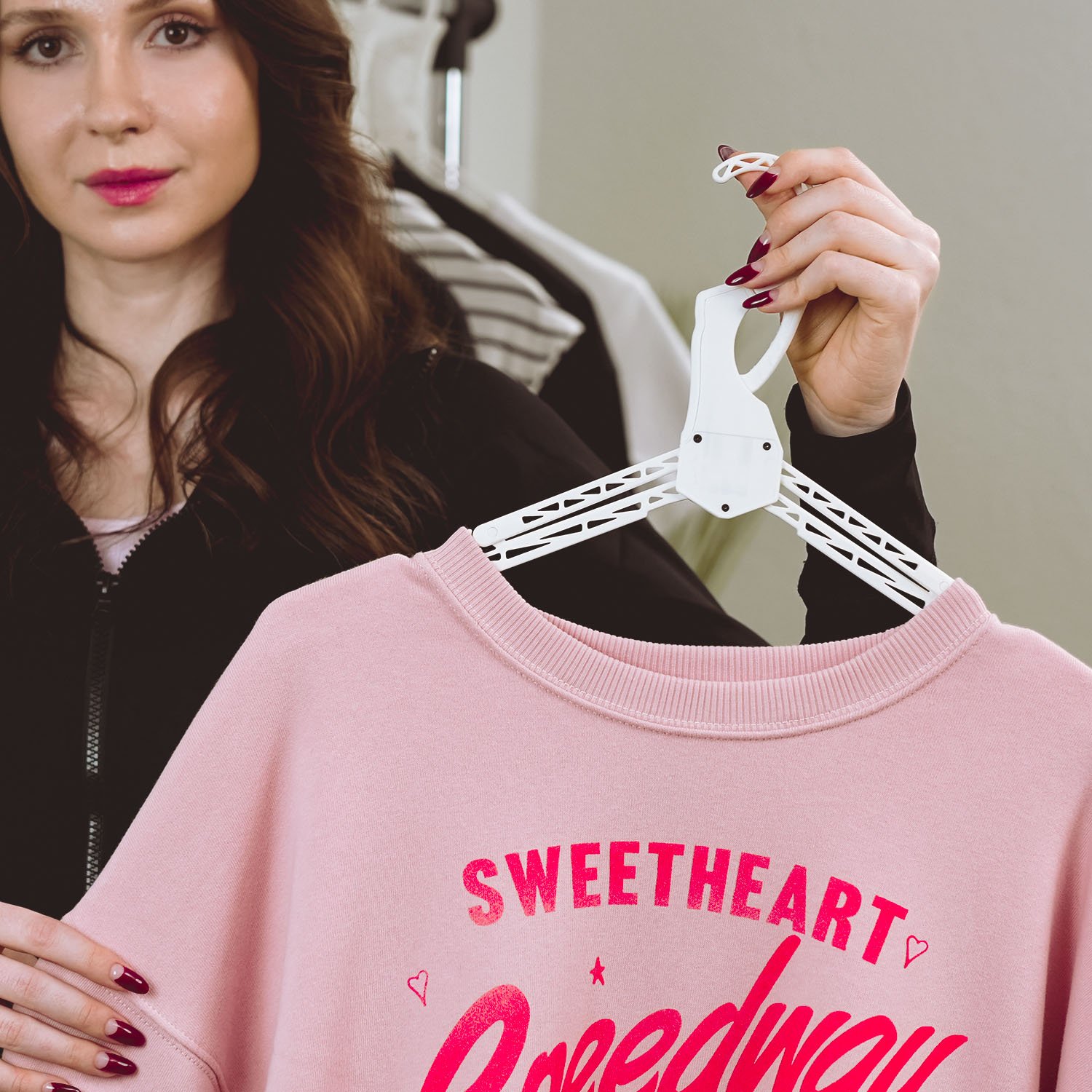 A woman with wavy brown hair and pink lipstick holding a white hanger with a pink sweatshirt that says 'SWEETHEART' in bold, pink letters and has small decorative hearts.